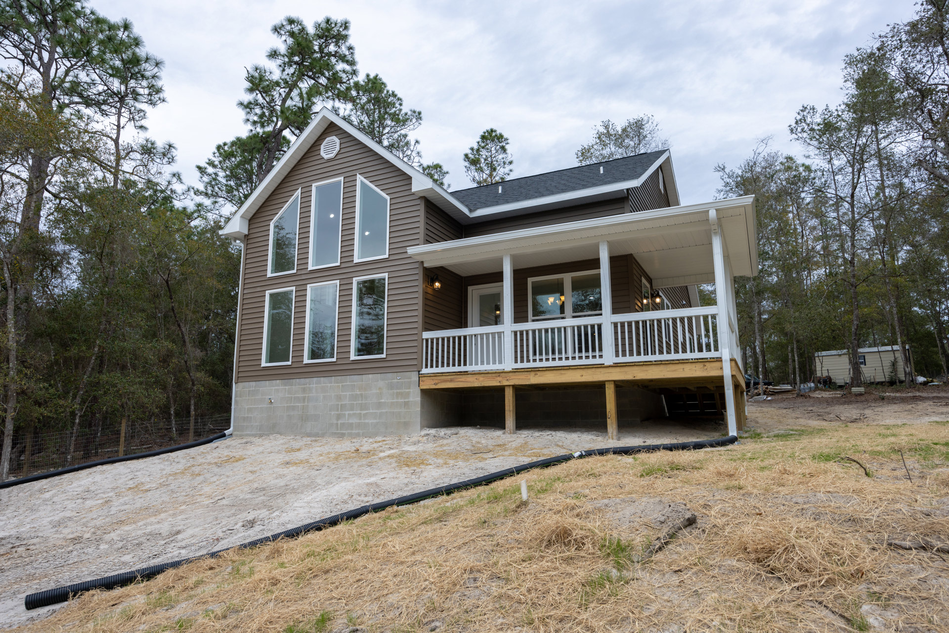 Two-story house with white siding, covered front porch, expansive wooden deck, multiple windows reflecting light, black pipe lying on the lawn, surrounded by trees and cloudy sky