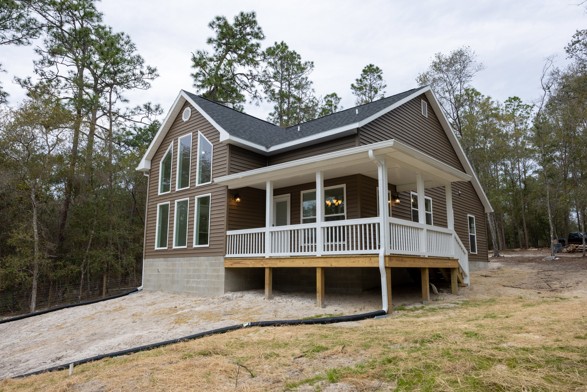Two-story house with white railing on wooden porch, expansive green lawn, mature trees in background, large windows, and light-colored siding