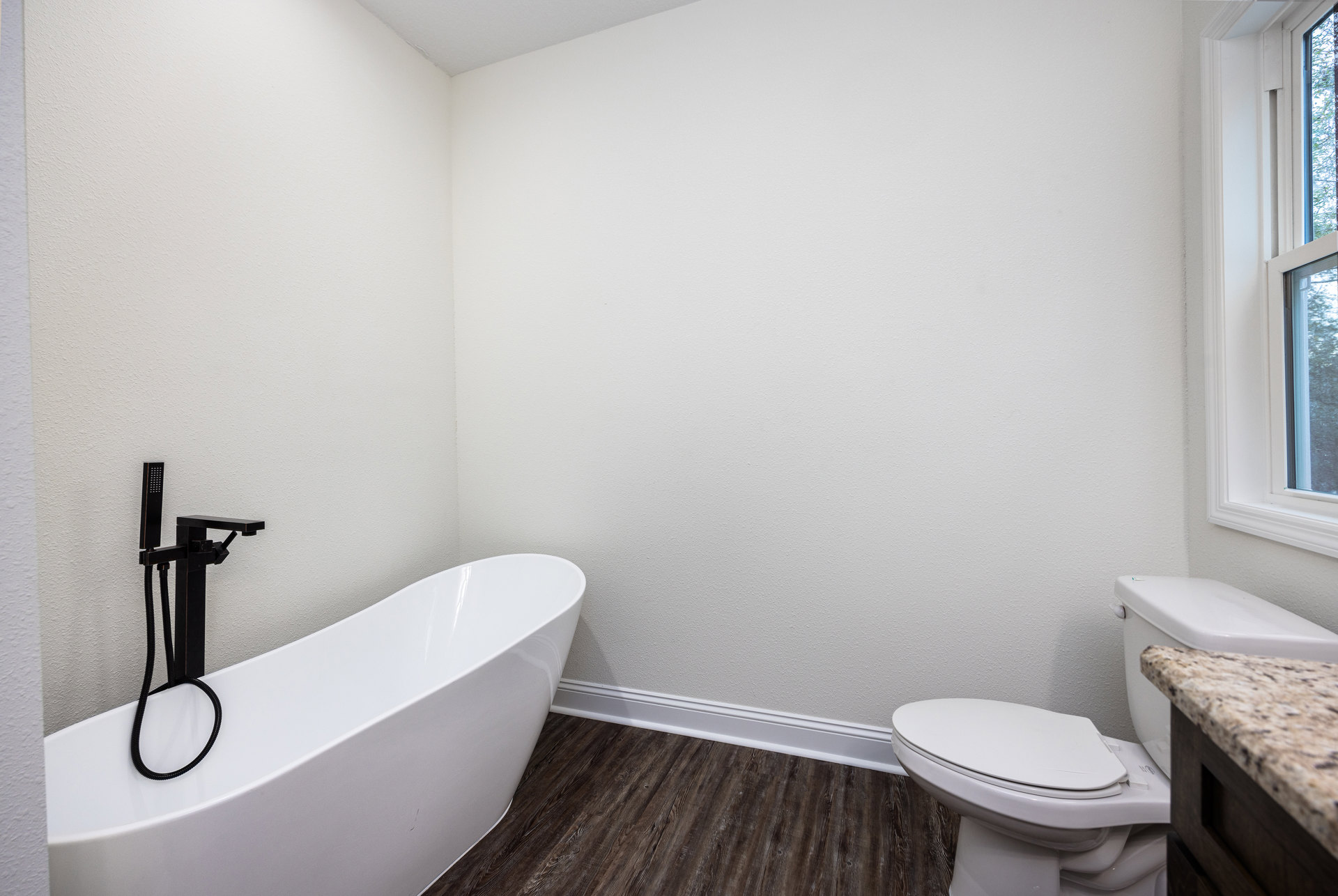 Bathroom with white bathtub featuring black hose and faucet, marble countertop with wood frame, toilet, and window with white frame; tile walls and floor.