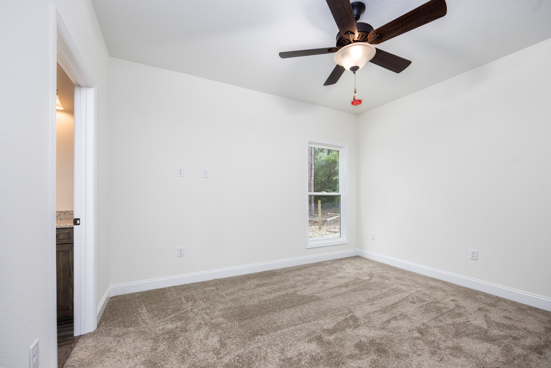 Neutral-toned carpeted bedroom with white walls, ceiling fan and light fixture, large window overlooking green trees