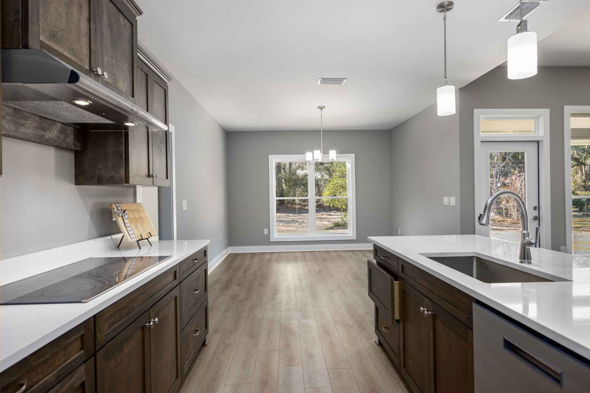 Dark wood kitchen cabinets, wood plank flooring, stone countertop with sink and faucet, window overlooking trees and bare ground, wood cutting board with ribbon, white appliance