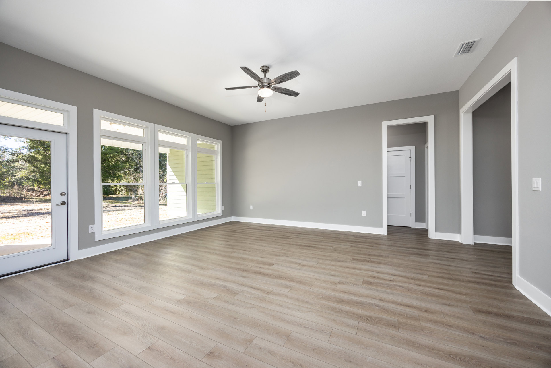 Ceiling fan with light fixture, wood laminate flooring, white walls, window with natural light, white door with black handle, ceiling vent