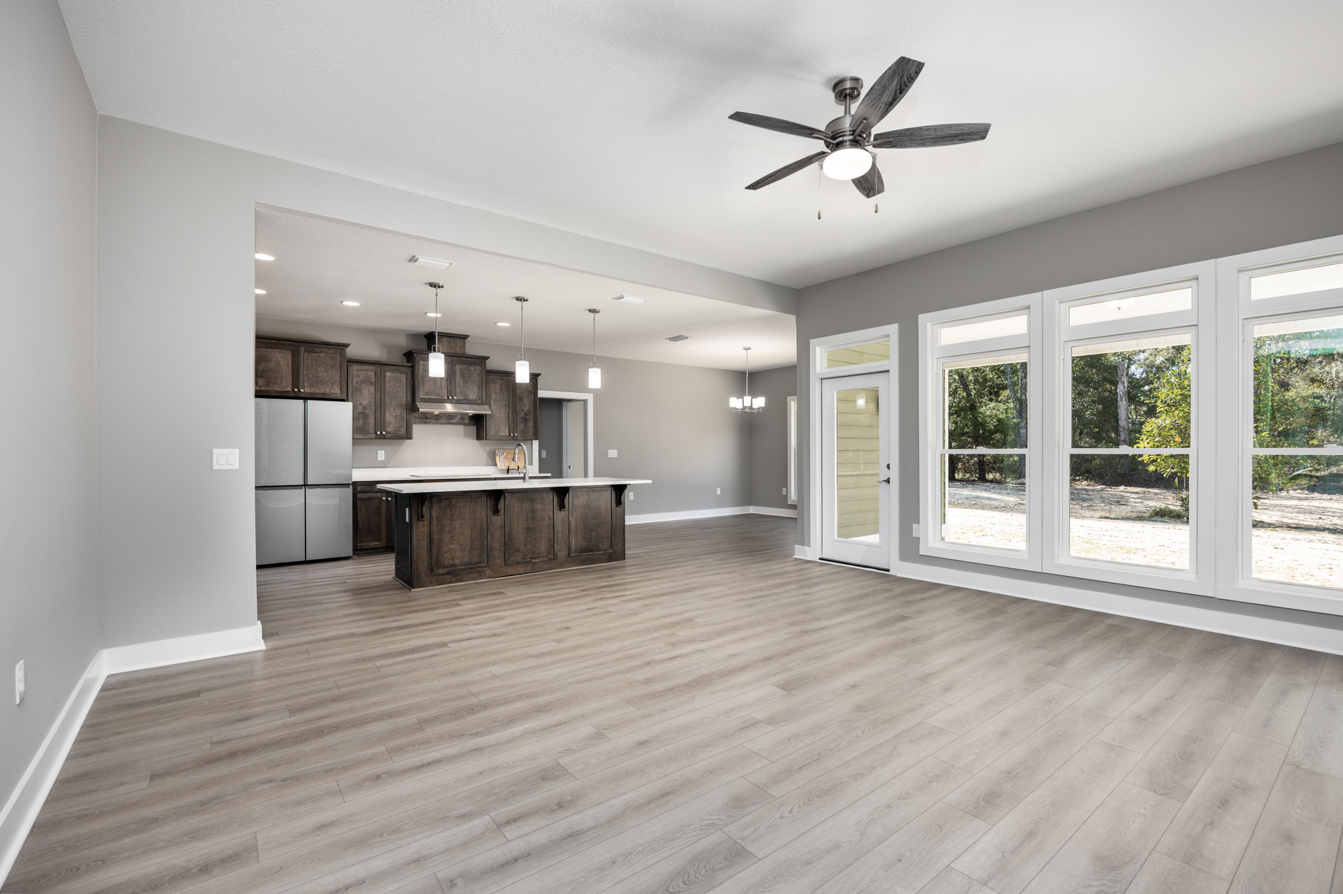 Spacious open floor plan featuring hardwood flooring, central kitchen island with sink and faucet, stainless steel refrigerator, adjacent dining area, and ceiling fan with light