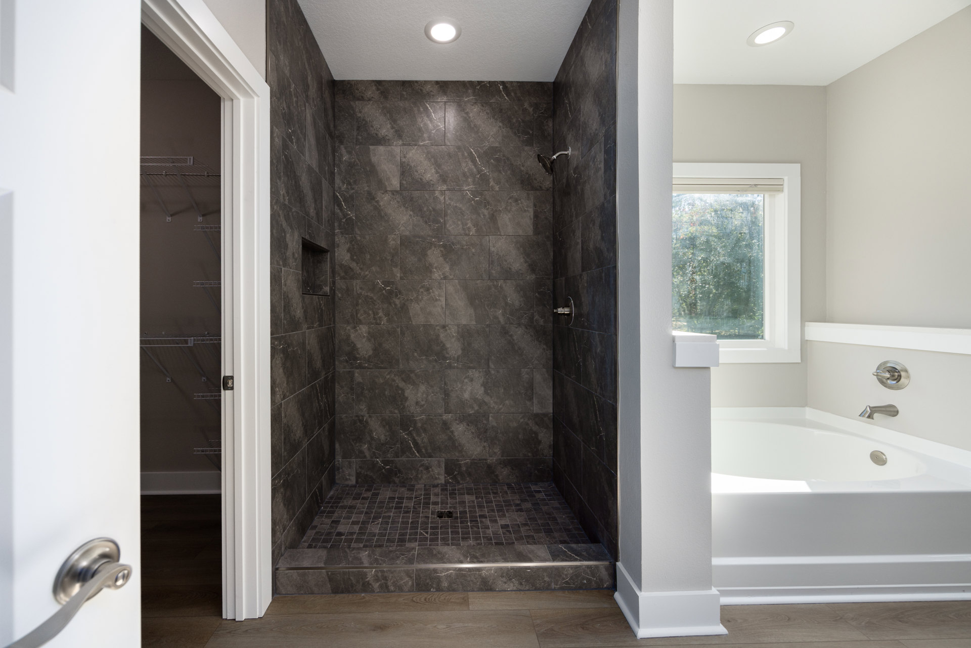 Bathroom featuring a glass-enclosed shower with gray tile floor, freestanding white bathtub with chrome faucet, large window overlooking trees, recessed lighting, and built-in