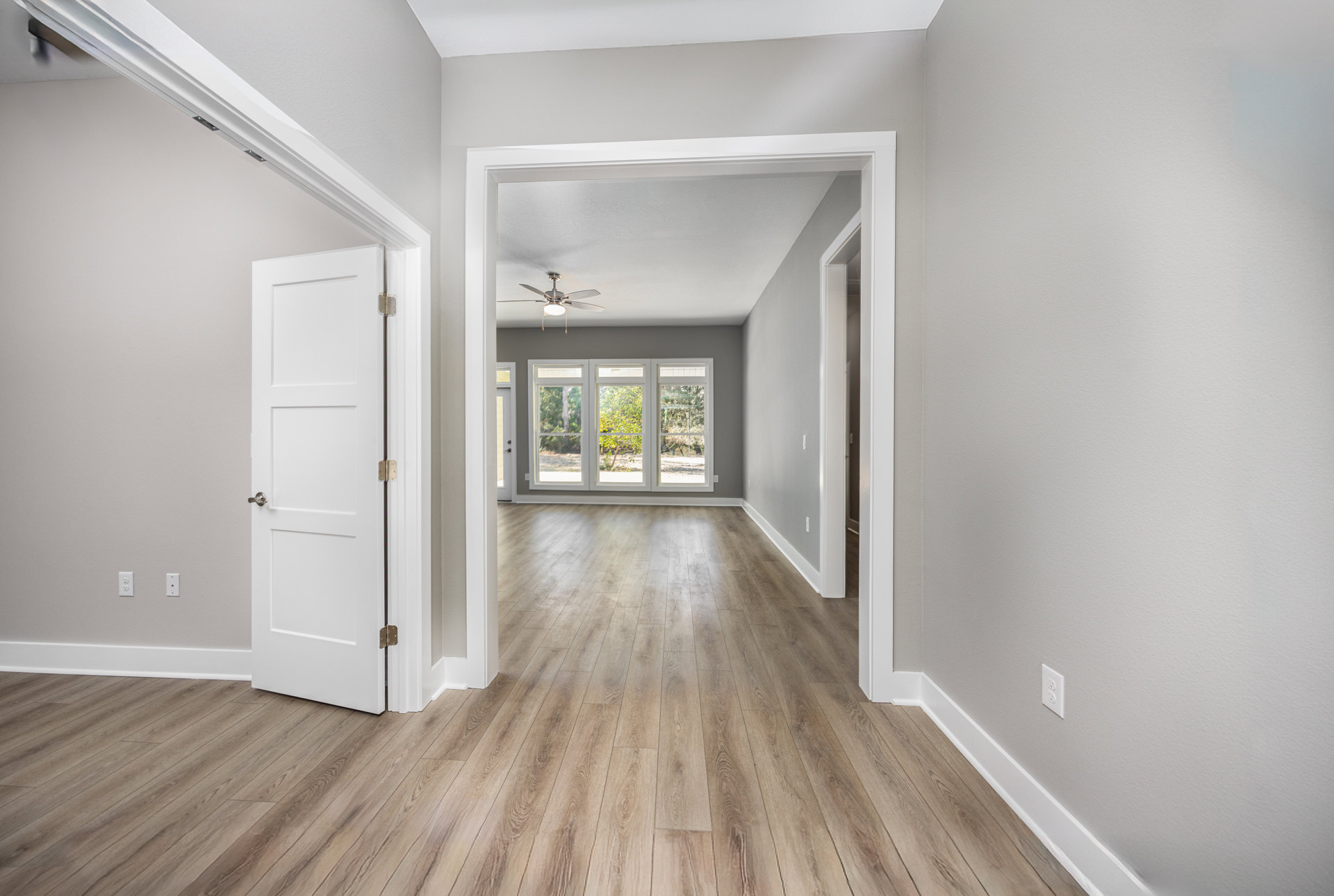 Hallway with wood flooring, white walls, ceiling fan with light, white door with silver handle, window showing green trees outside
