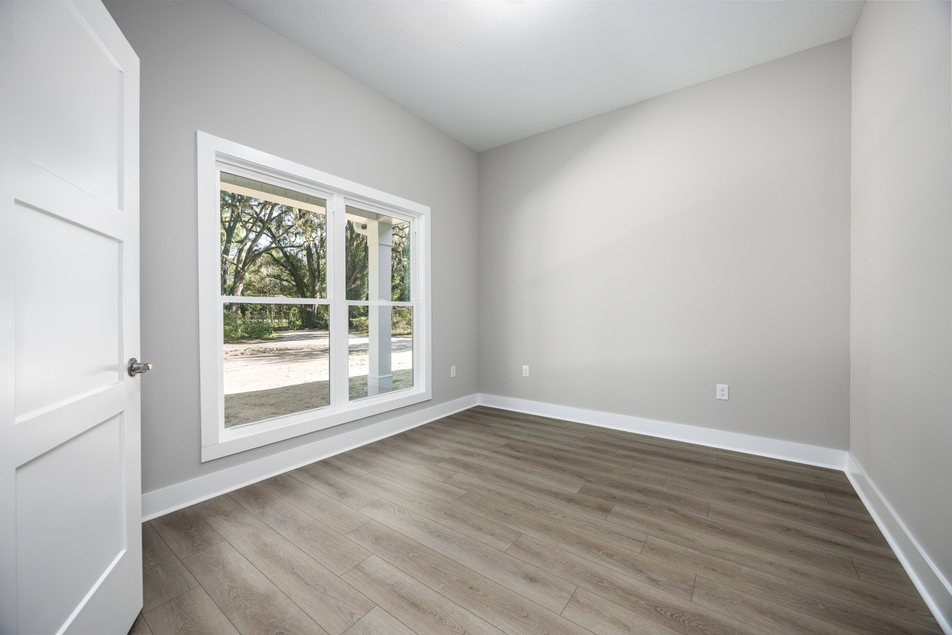 Hardwood floor with white walls, large window showing trees outside, white door with silver handle