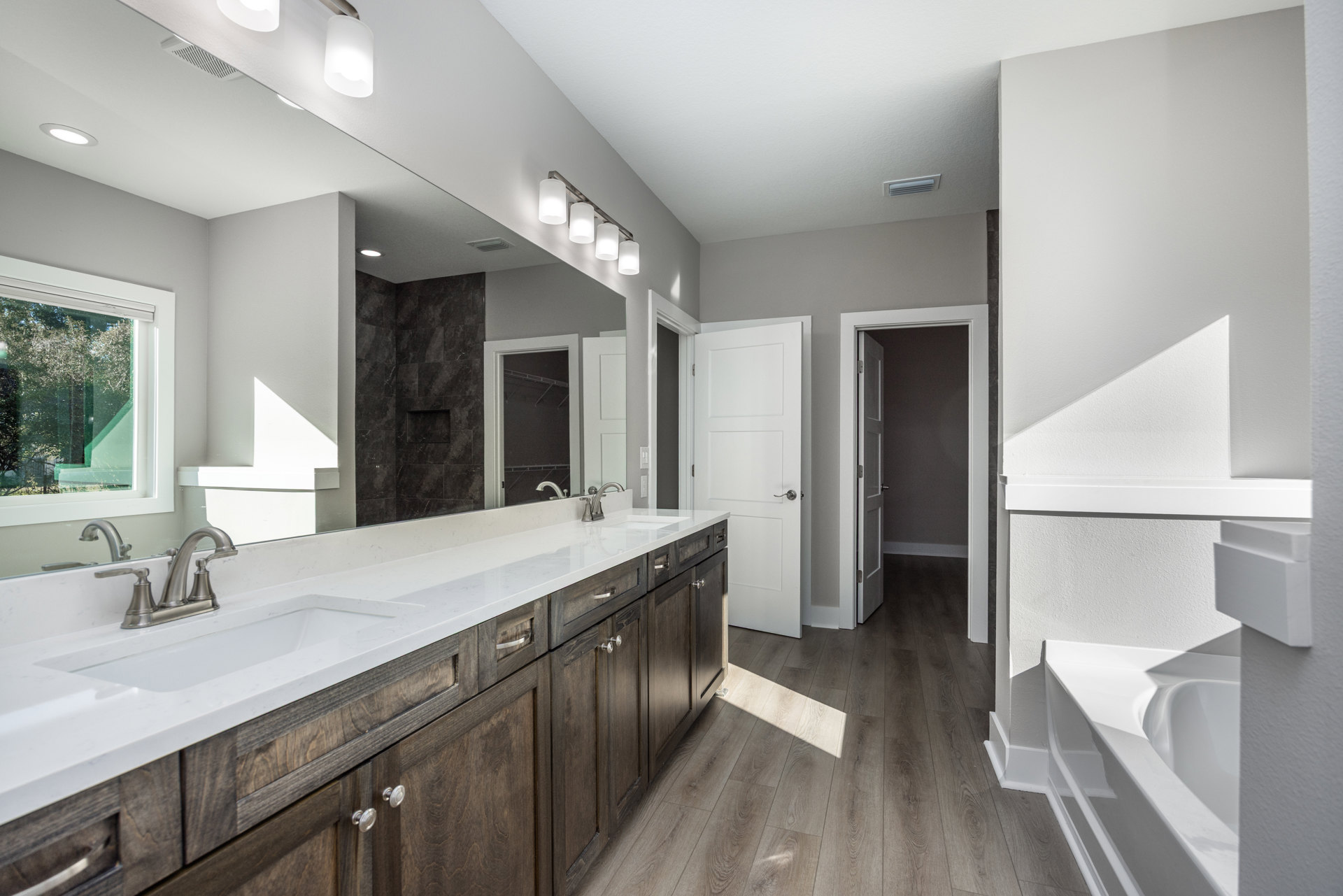 Bathroom with a wide mirror above a rectangular sink, white cabinetry, silver faucet, four-light fixture, window showing trees, and a white door with silver handle.