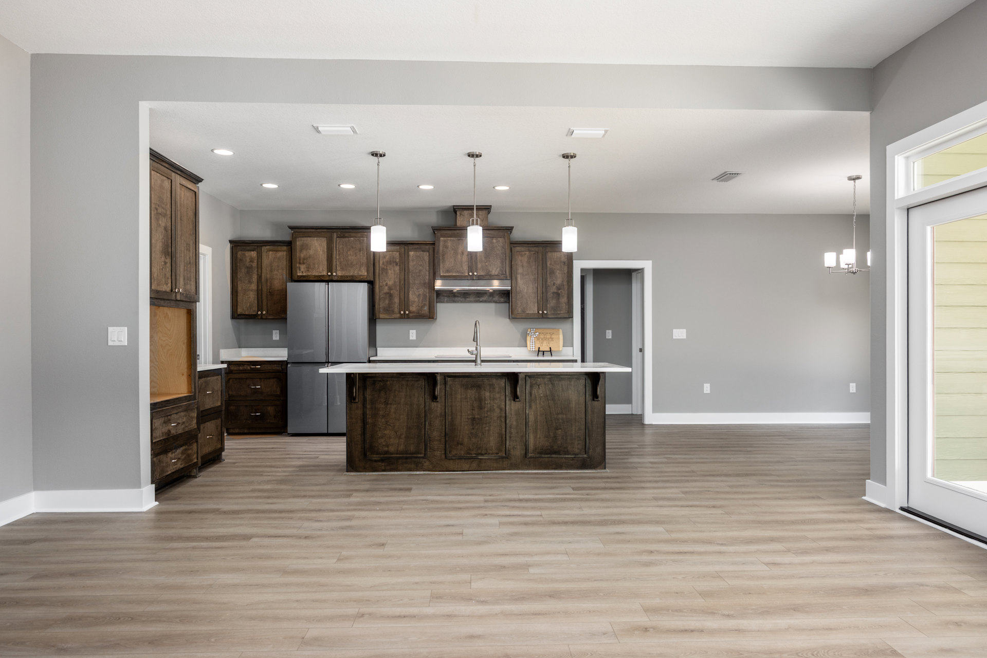 Spacious kitchen featuring a large central island with white countertop, stainless refrigerator, undermount sink, wood flooring, white cabinetry, tile backsplash, and a window near