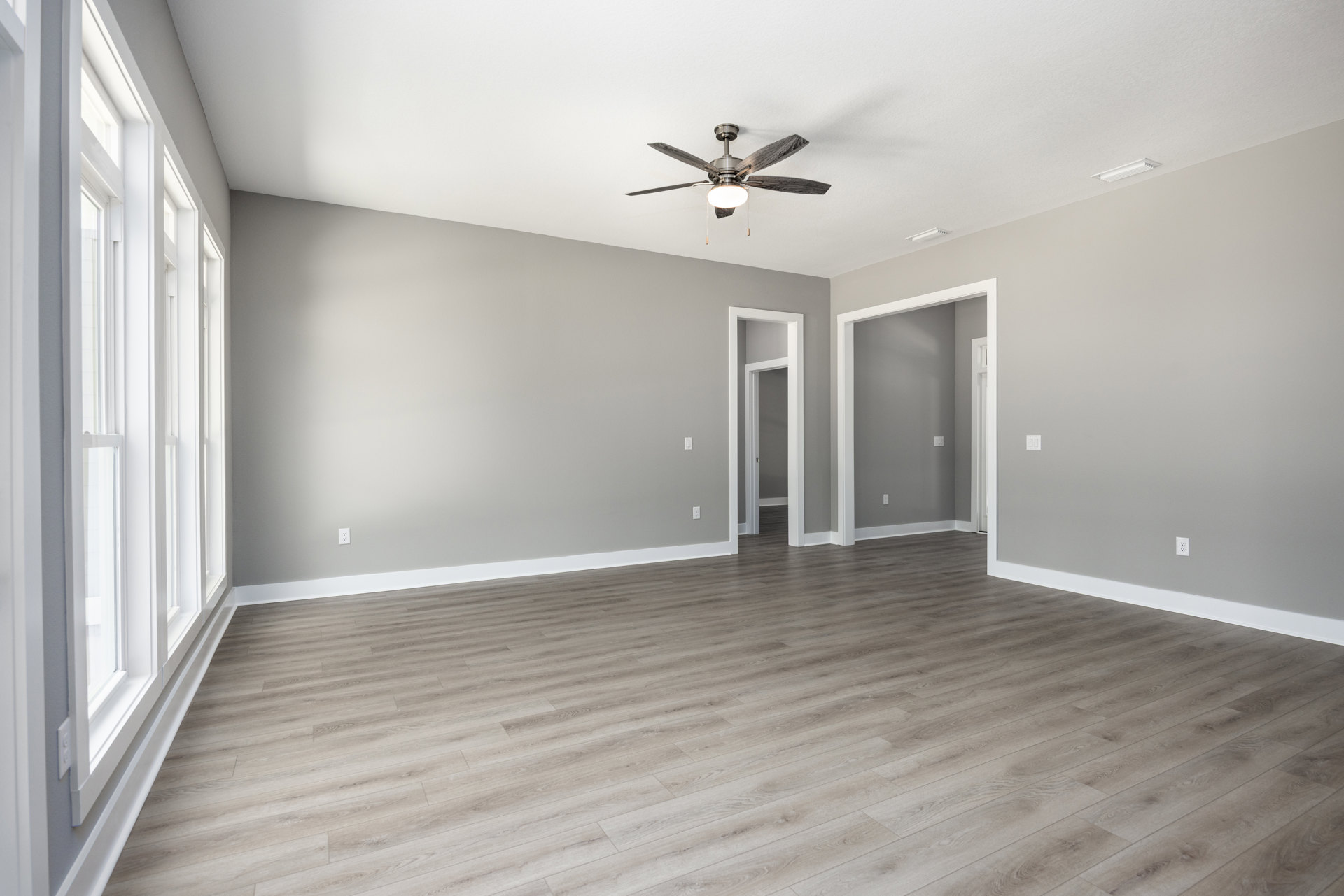 Ceiling fan with light fixture above wood flooring, white baseboards, multiple windows with white frames, door frame featuring light switch, neutral plaster walls