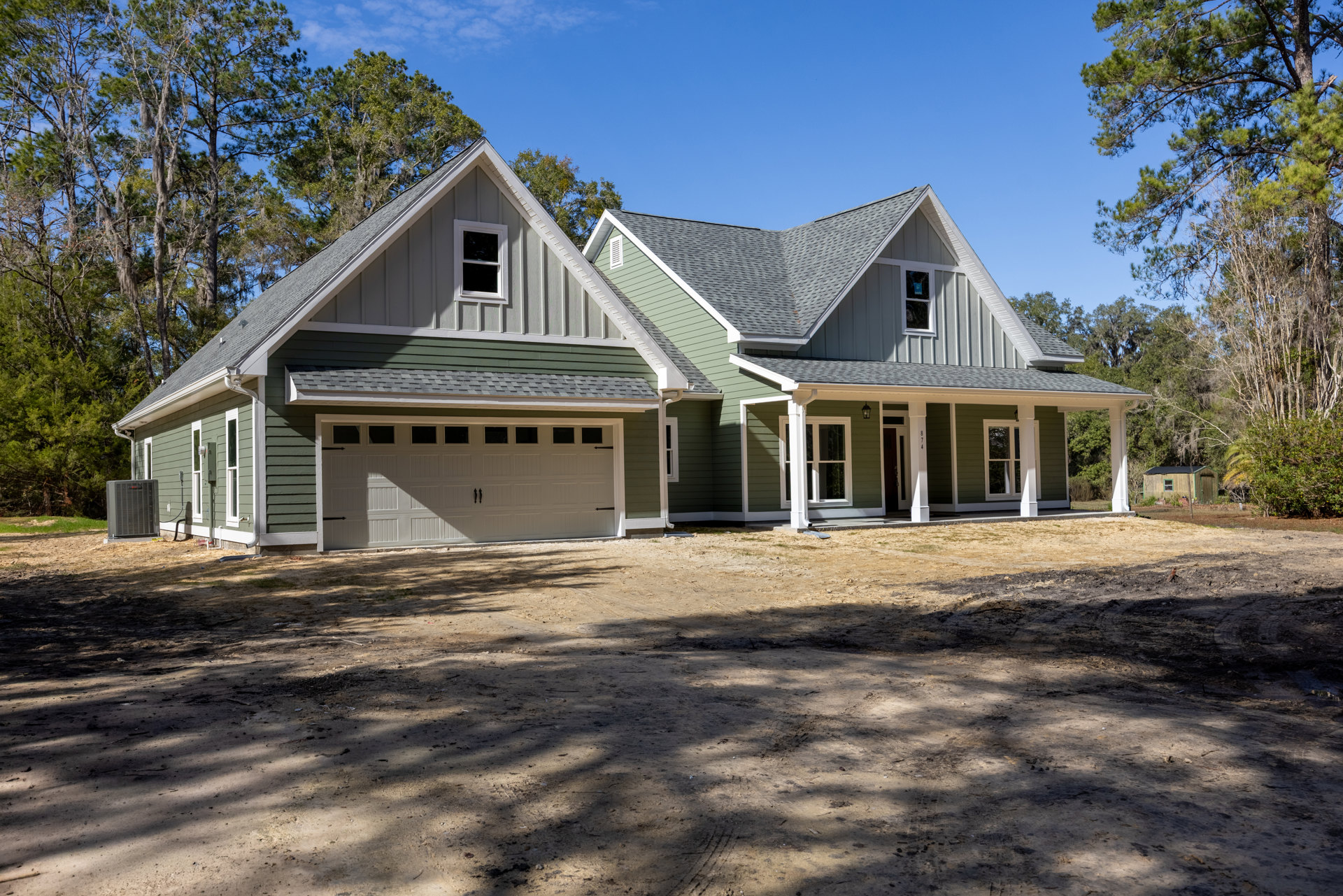 White garage door on a house with broken window glass, dirt driveway, grey outdoor heater, and trees lining the background