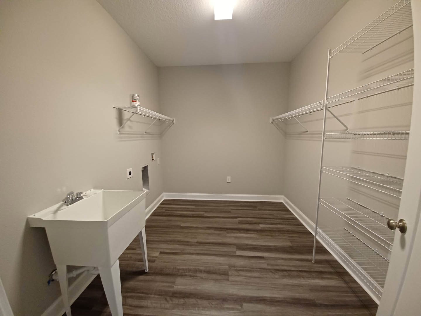 White porcelain sink with chrome faucet and exposed legs, wood flooring with white baseboards, white metal shelving mounted on plaster wall, close-up of brushed metal door knob.