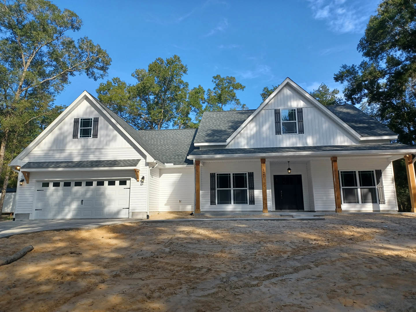 White siding house with gray roof, black front door, white-framed window, dirt driveway with tire tracks, surrounded by trees in the background