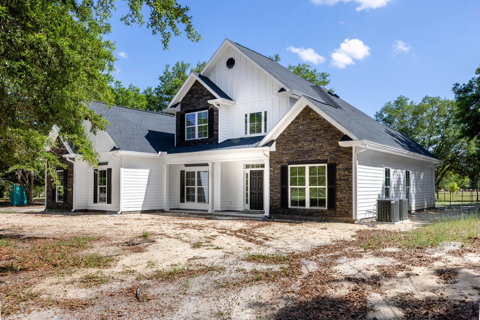 Two-story house with brick and stone exterior, white-framed windows, surrounded by dirt patches and mature trees, blue portable toilet near wooded area