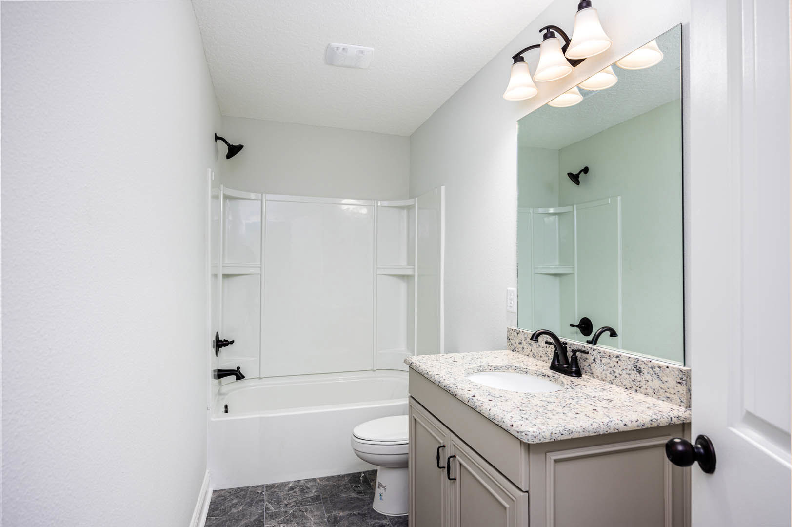 Bathroom with white tile walls, black faucet on rectangular sink, wall-mounted mirror, multi-bulb light fixture above, closed toilet, vented white object, and black shower head