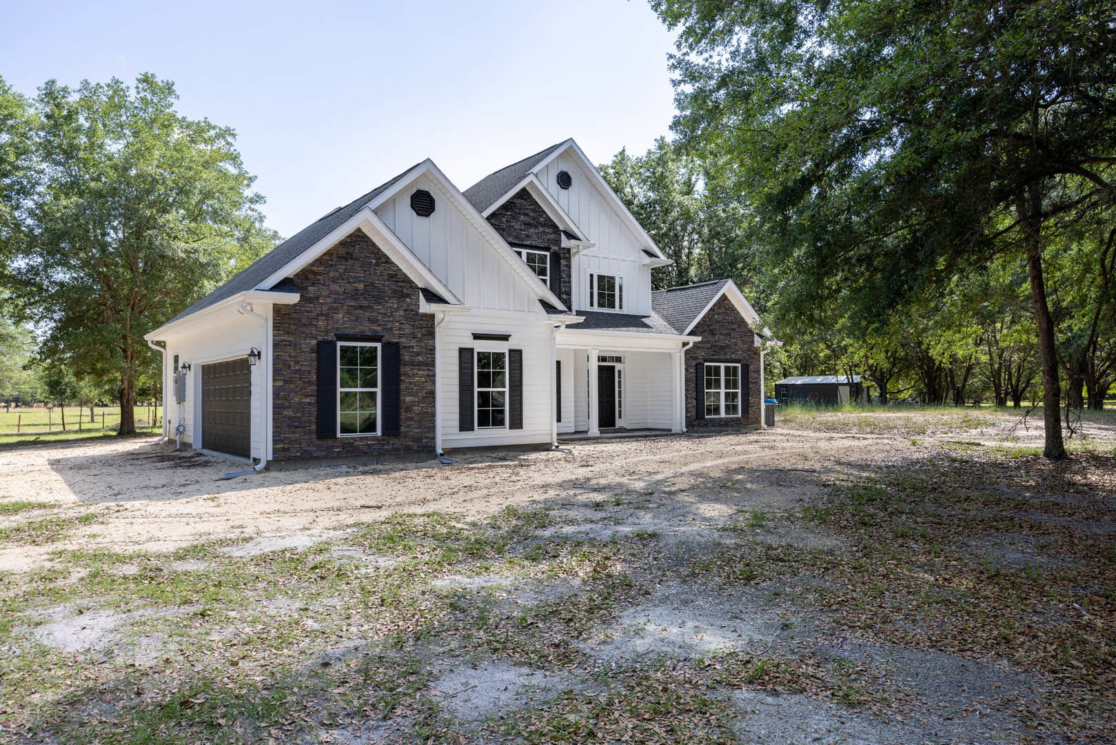 Brick exterior home with white and black roof, black shuttered windows, dirt driveway, and mature trees in the background