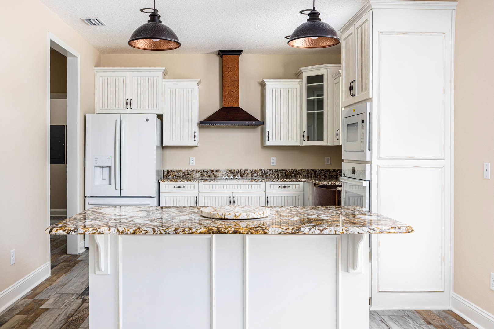 Marble countertops and white cabinets in a kitchen, white refrigerator with ice and water dispenser, circular object on countertop, ceiling light fixture, view into long hallway