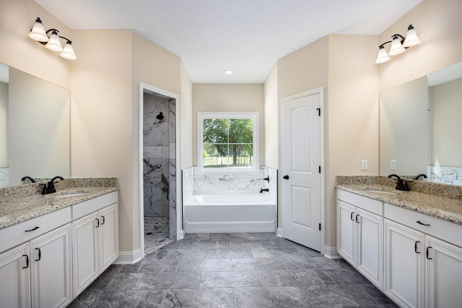 Bathroom featuring marble countertops, freestanding tub, grey tile floor with white grout lines, white door with black knobs, window overlooking trees, three-lamp light fixture