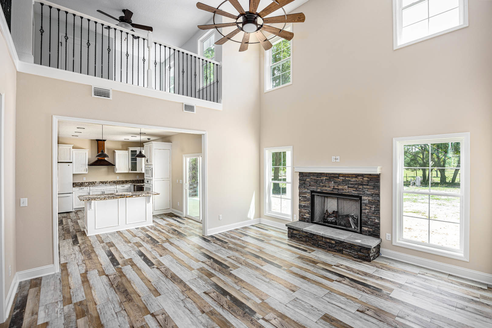 Living room with hardwood floor, stone fireplace, ceiling fan with light fixture, large window overlooking trees and grass, person seated in window nook, adjacent white kitchen