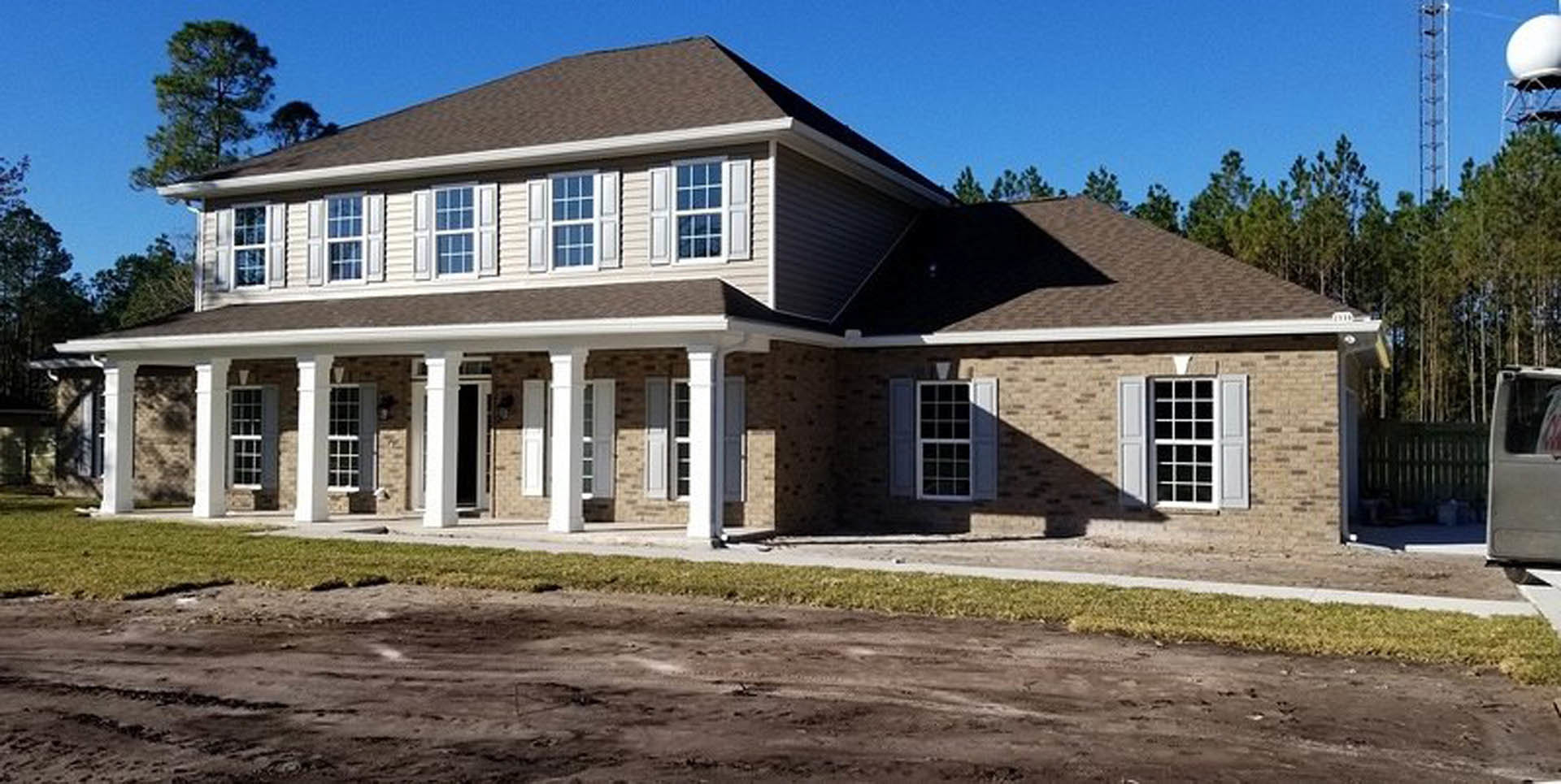 Two-story house with wide front porch, white columns, light-colored siding, multiple windows with white frames and shutters, dirt driveway bordered by grass, blue sky overhead