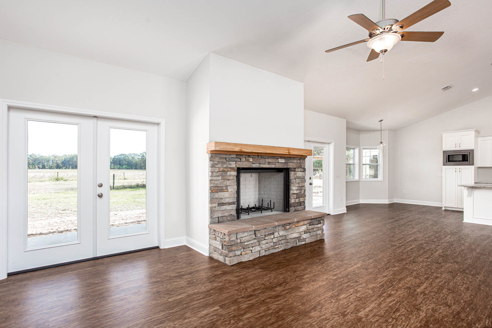 Living room with wood flooring, stone fireplace, ceiling fan with light fixture, double glass-paneled doors, and microwave oven visible in adjacent space