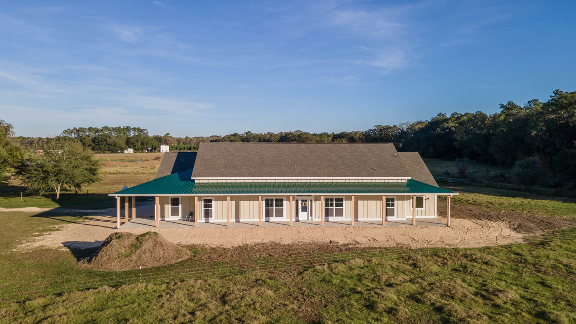 Green metal roof atop a cottage-style home, multi-pane windows, wooden fence in foreground, leafy tree, grassy yard, blue sky with scattered clouds