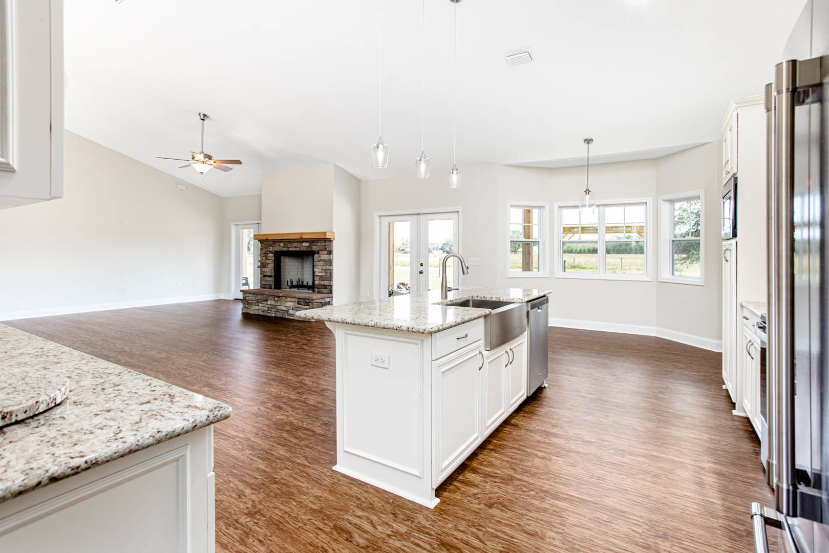 Open-concept kitchen and living room featuring a stone fireplace, white cabinetry, quartz countertops, stainless steel appliances, kitchen island with built-in sink, hardwood