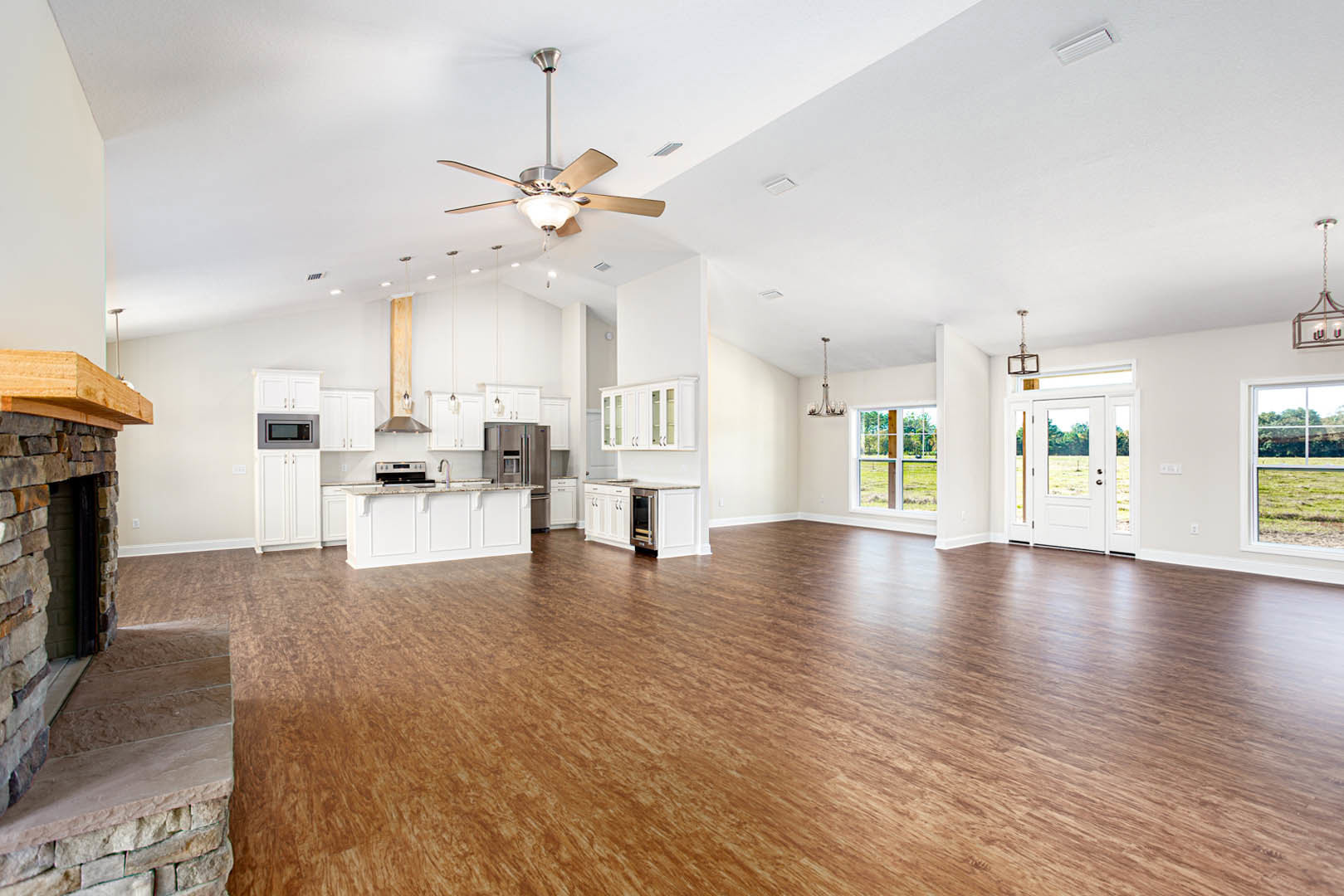 Spacious open floor plan featuring hardwood flooring, stone accent wall, ceiling fan with light fixture, kitchen counter near glass door, and white door with glass panes