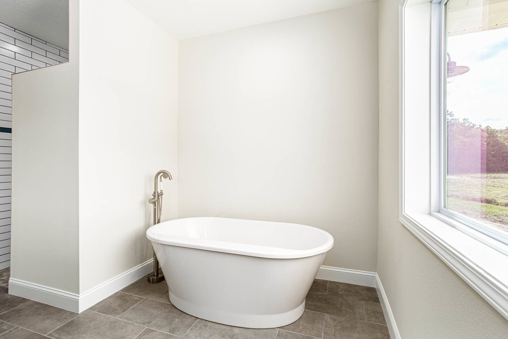 Freestanding white bathtub with chrome faucet, light gray tile floor, large window overlooking trees, white walls, and modern bathroom fixtures