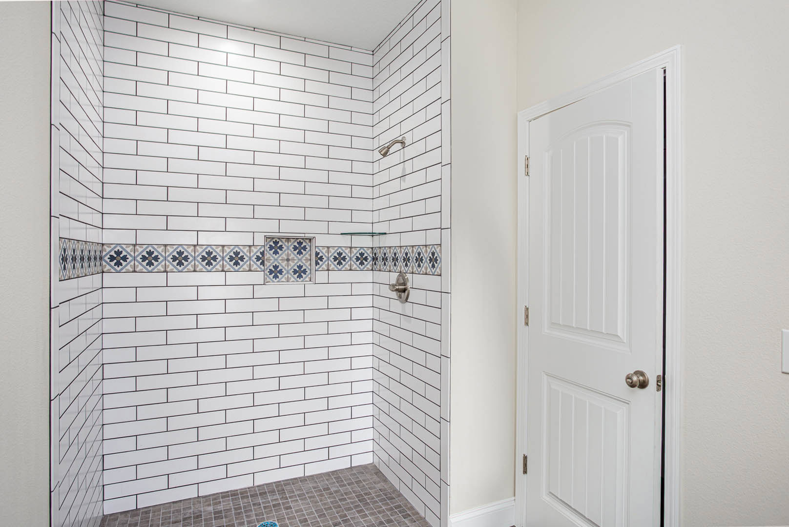 Bathroom with white tile walls and black grout, glass shower enclosure, white door with silver handle, and light gray tile flooring