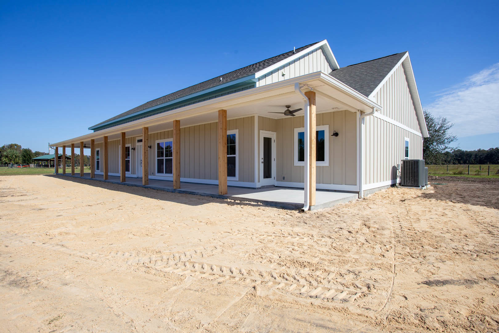 White custom home with prominent pillars, covered porch featuring ceiling fan, unfinished dirt yard with visible tire tracks, large windows, and dark front door under a gabled