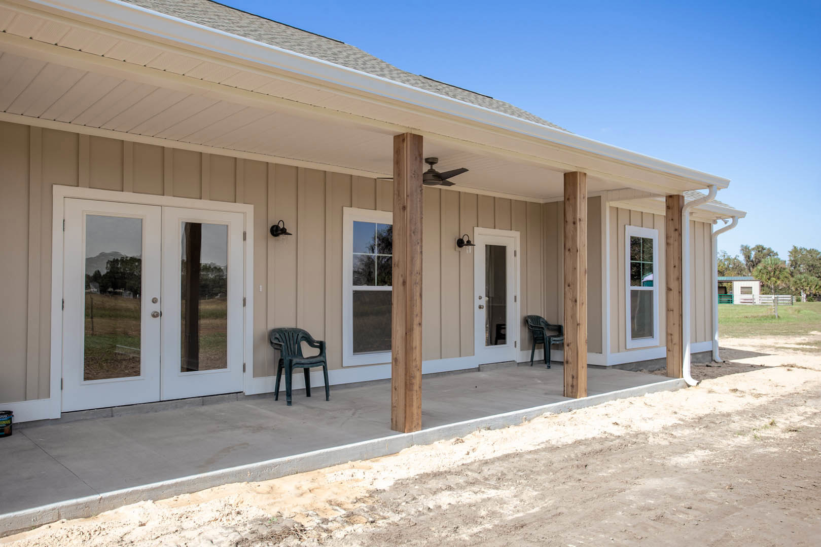 Covered porch with white ceiling, glass double doors, white-framed window, plastic chairs, and dirt driveway beside the house