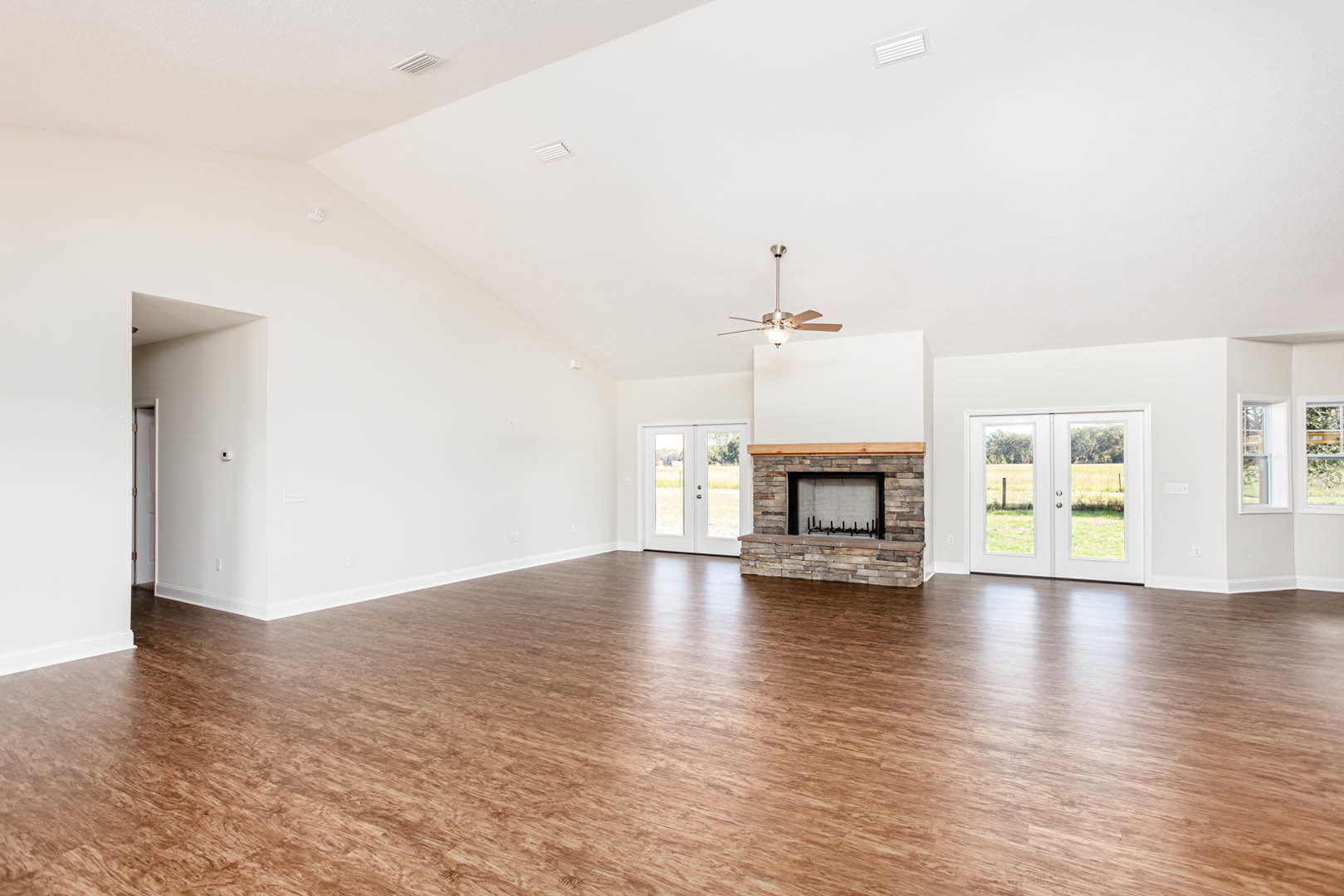 Spacious living room with hardwood floors, stone fireplace, glass-paneled double doors, ceiling fan with light, and white walls