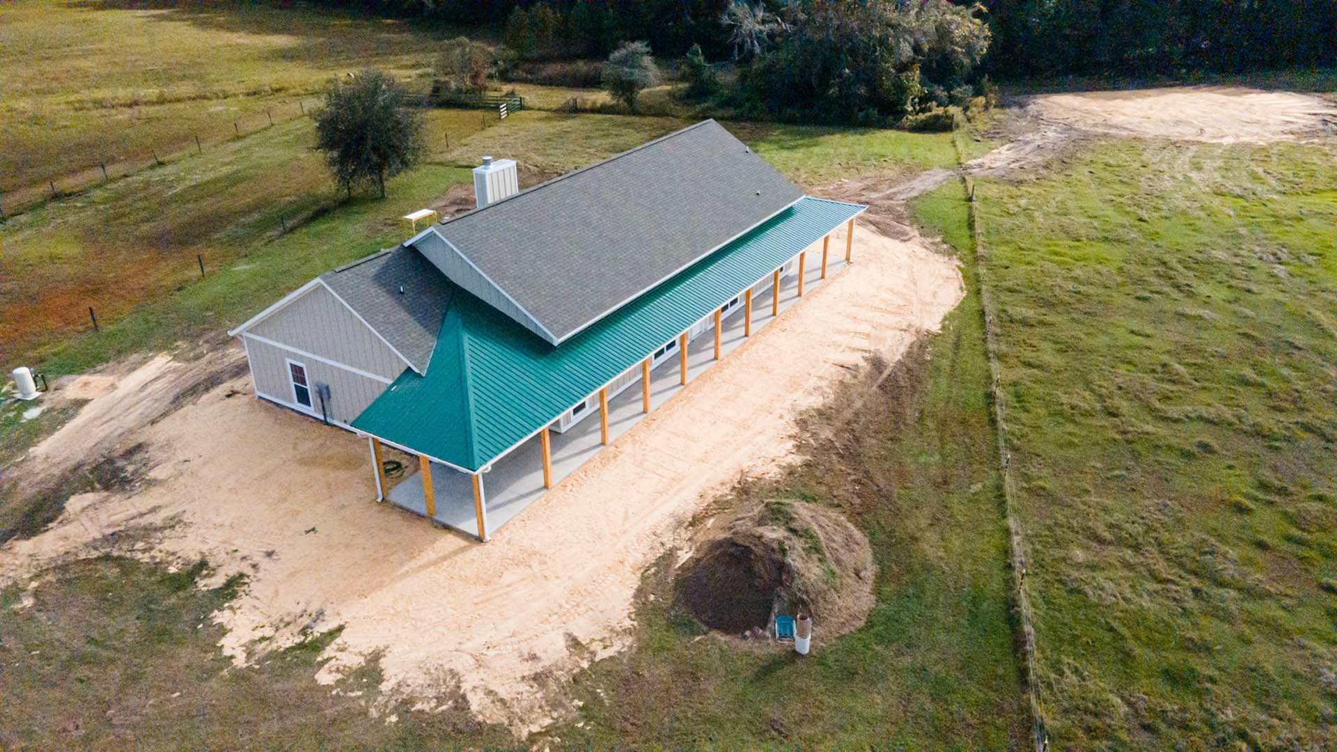 Partially built house with exposed foundation, green metal roof, white vent pipe, surrounding grassy lot, wooden fence, and nearby bushes and trees