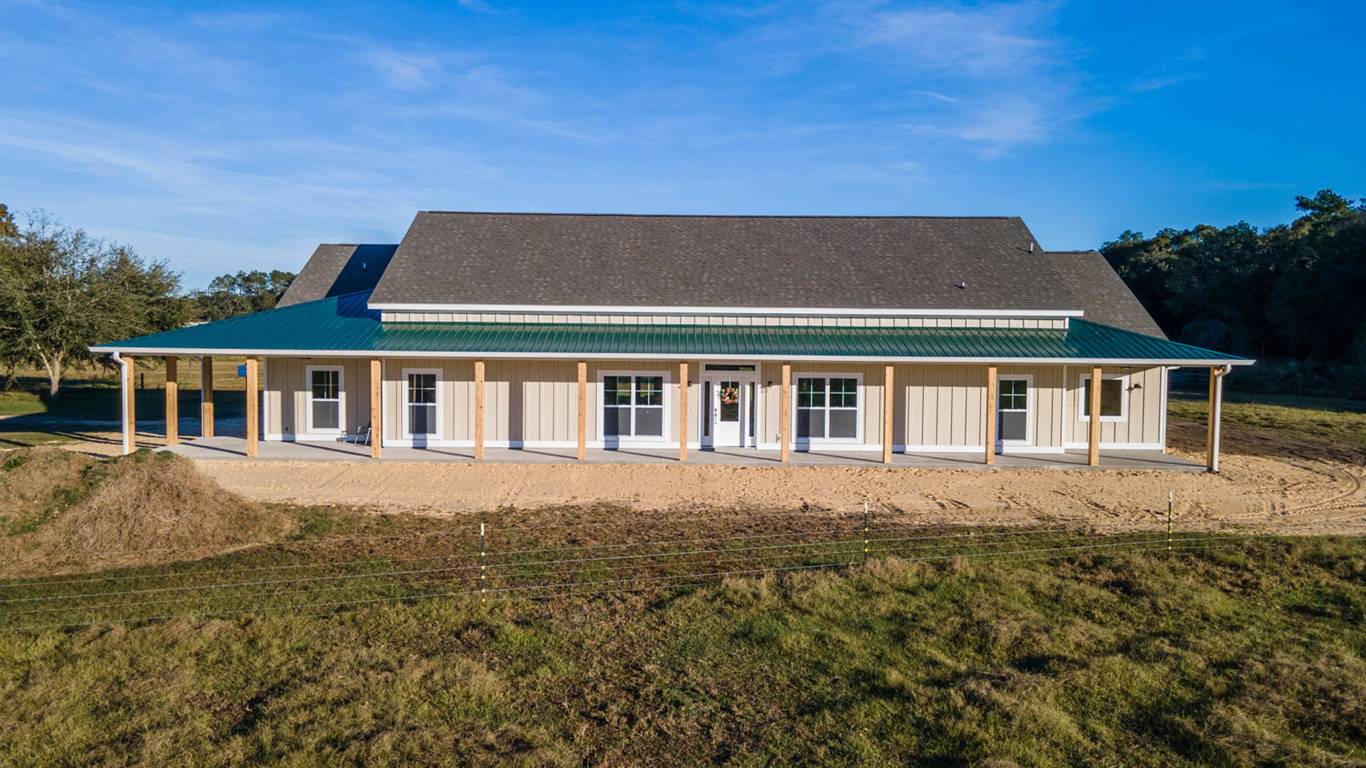 Green metal roof atop a cottage-style home, white-framed window, white front door with hanging basket, wire fence bordered by grass, blue sky and scattered clouds in background.