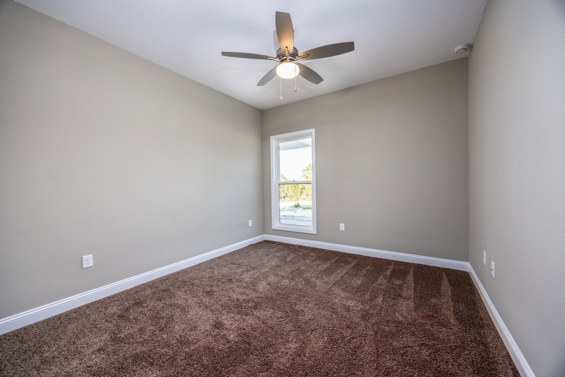 Ceiling fan with central light fixture, beige carpet flooring, white walls, large window overlooking green trees