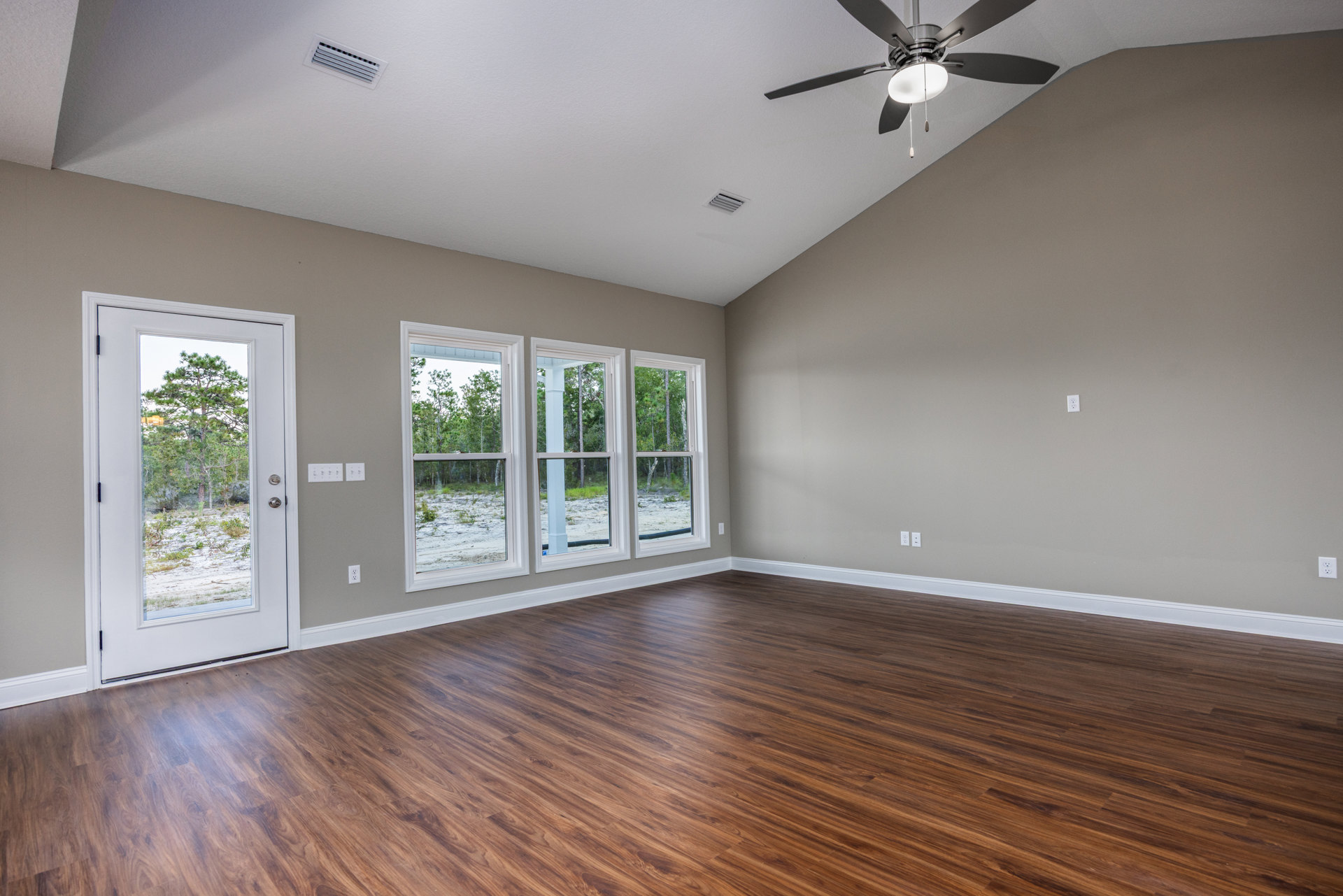 Wood flooring in a bright room with a ceiling fan, multiple windows showing trees and snow outside, and a door opening to a snowy landscape.