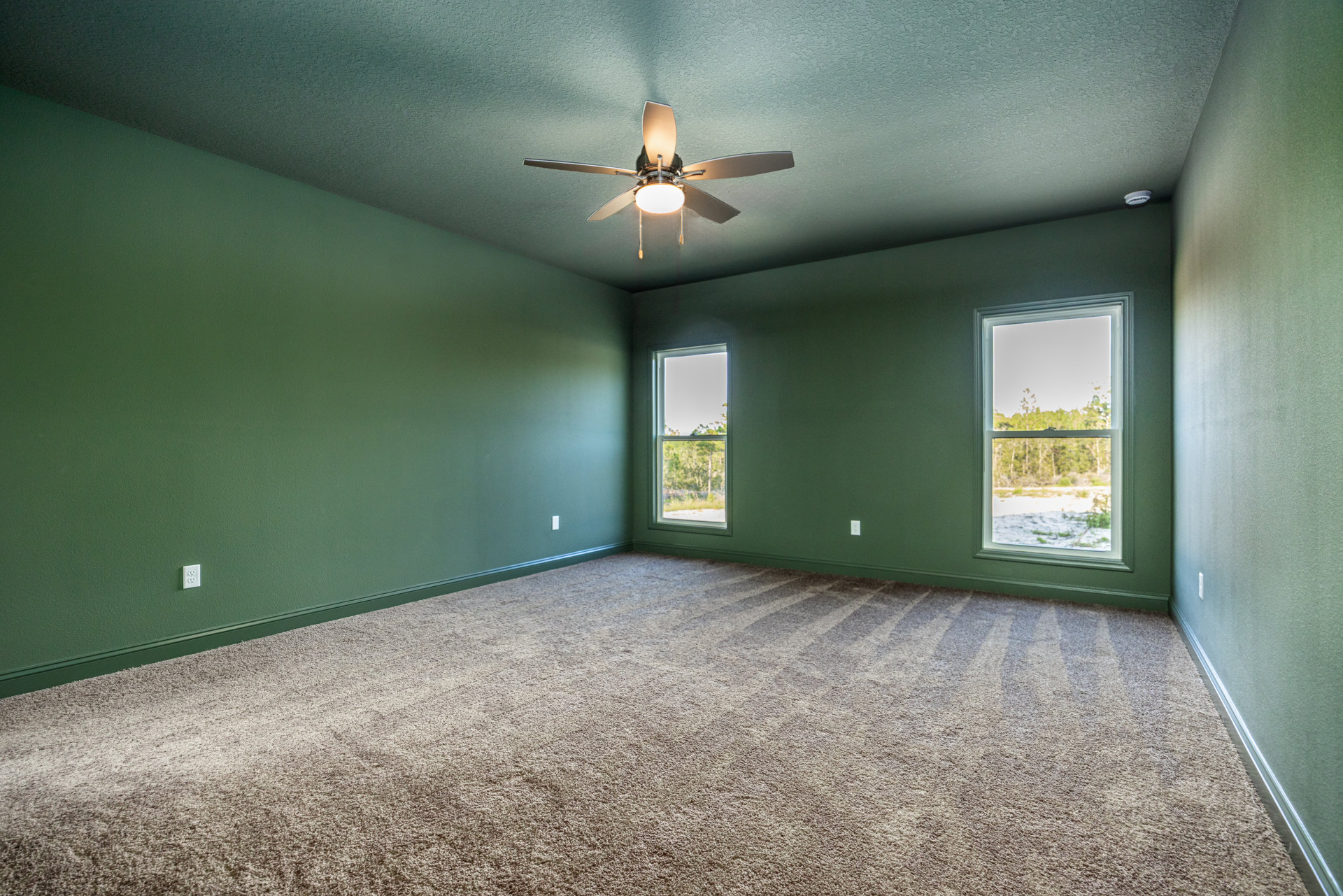 Carpeted room with white plaster walls, ceiling fan with light fixture, large windows showing green trees outside