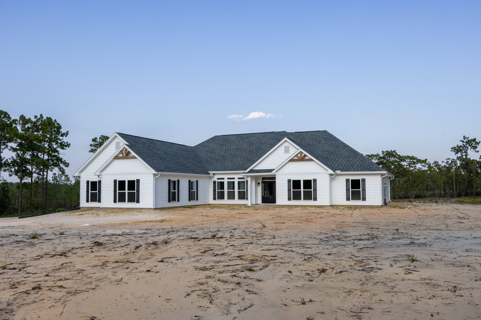 Modern house with black front door, light-colored siding, and pitched roof, surrounded by dirt yard with scattered plants and bordered by dense trees.