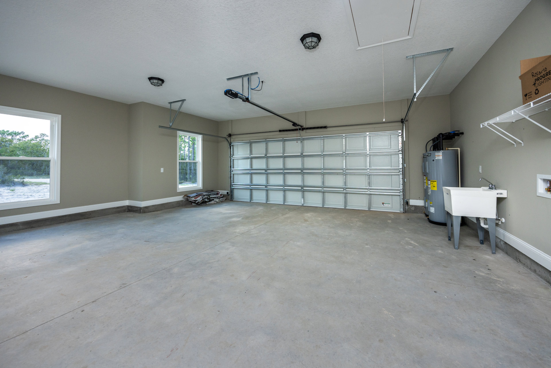 Spacious garage featuring a wide white overhead door, freestanding white utility sink with silver faucet, wall-mounted light fixture, large grey storage container with yellow