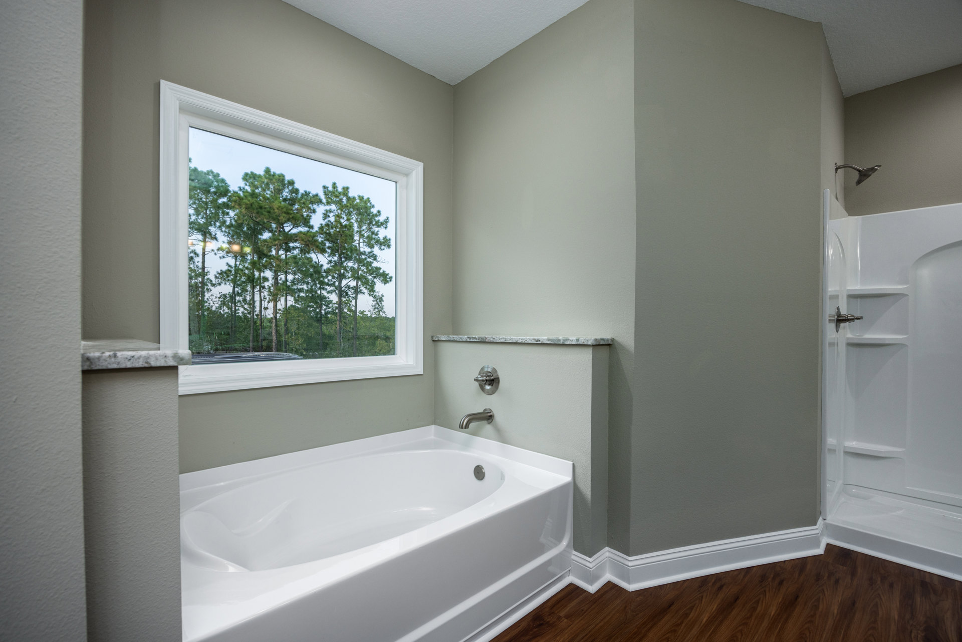 White freestanding bathtub with chrome faucet beside a window showing green trees, adjacent to a tiled shower stall with glass door and curved shower head, bathroom walls finished