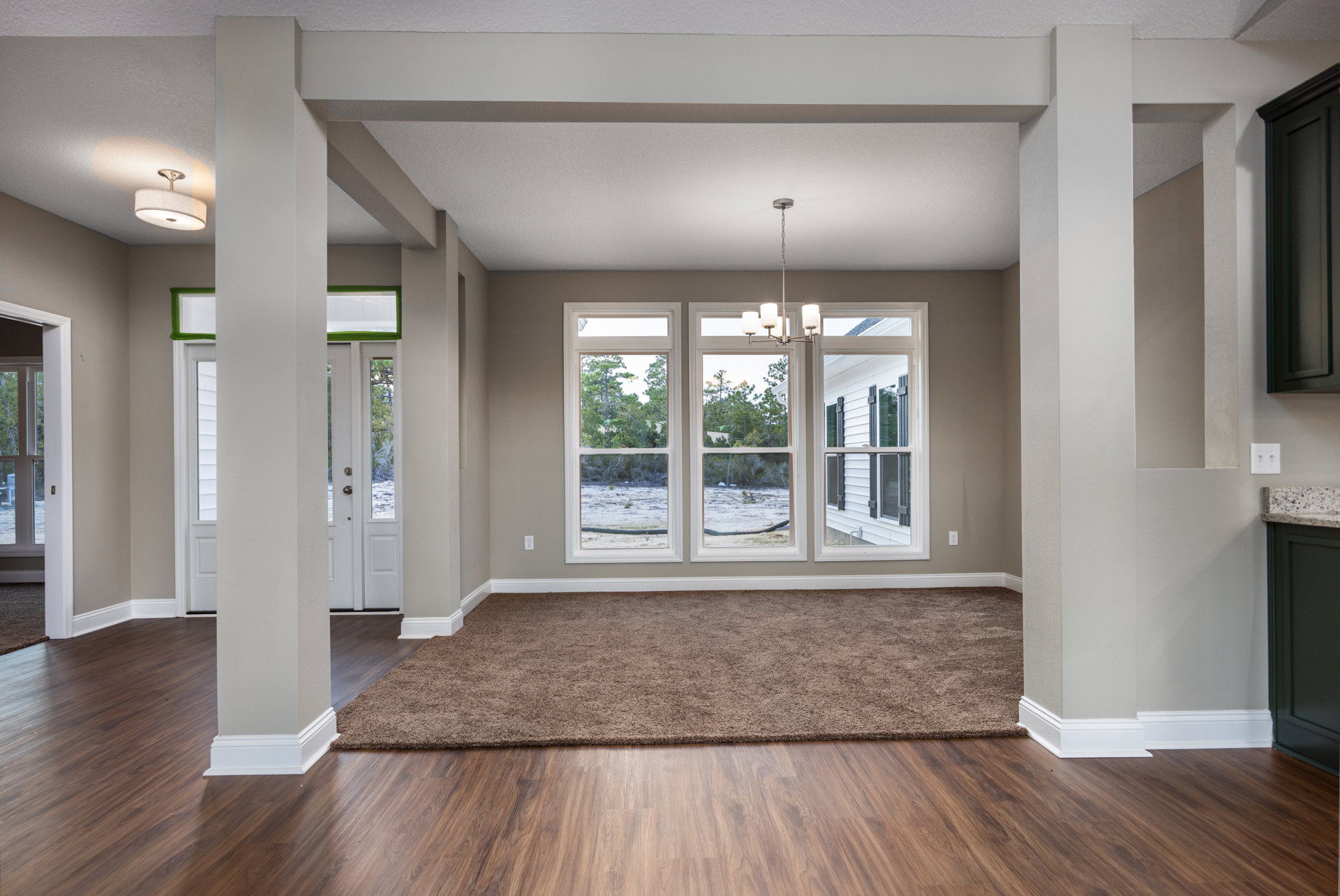 Brown carpet covering part of a wood floor in a room with a white ceiling, large window showing trees outside, and a chandelier hanging near the window.