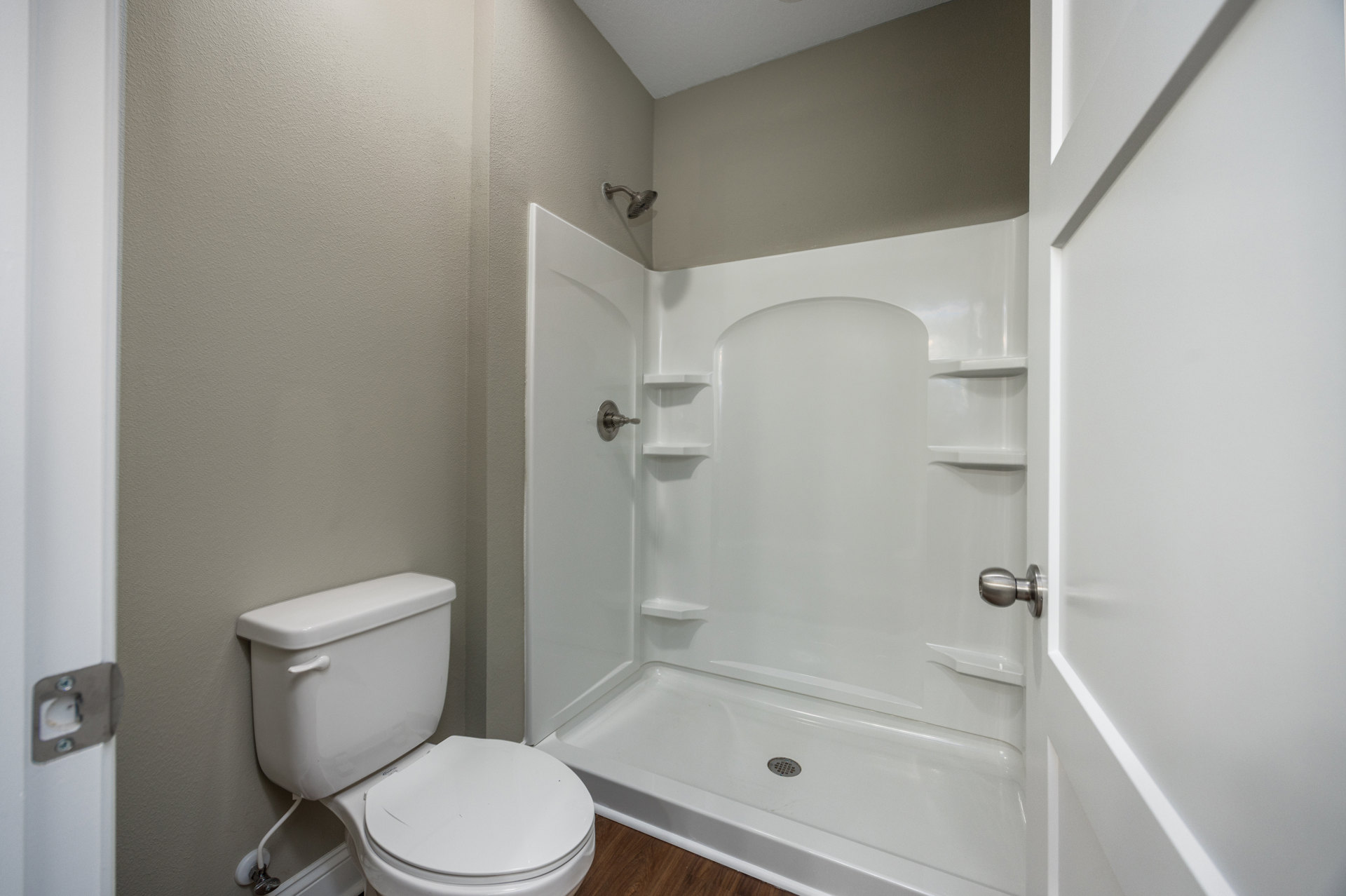 White toilet with open lid beside a tiled shower enclosure, chrome showerhead, and wall outlet on light-colored bathroom walls.