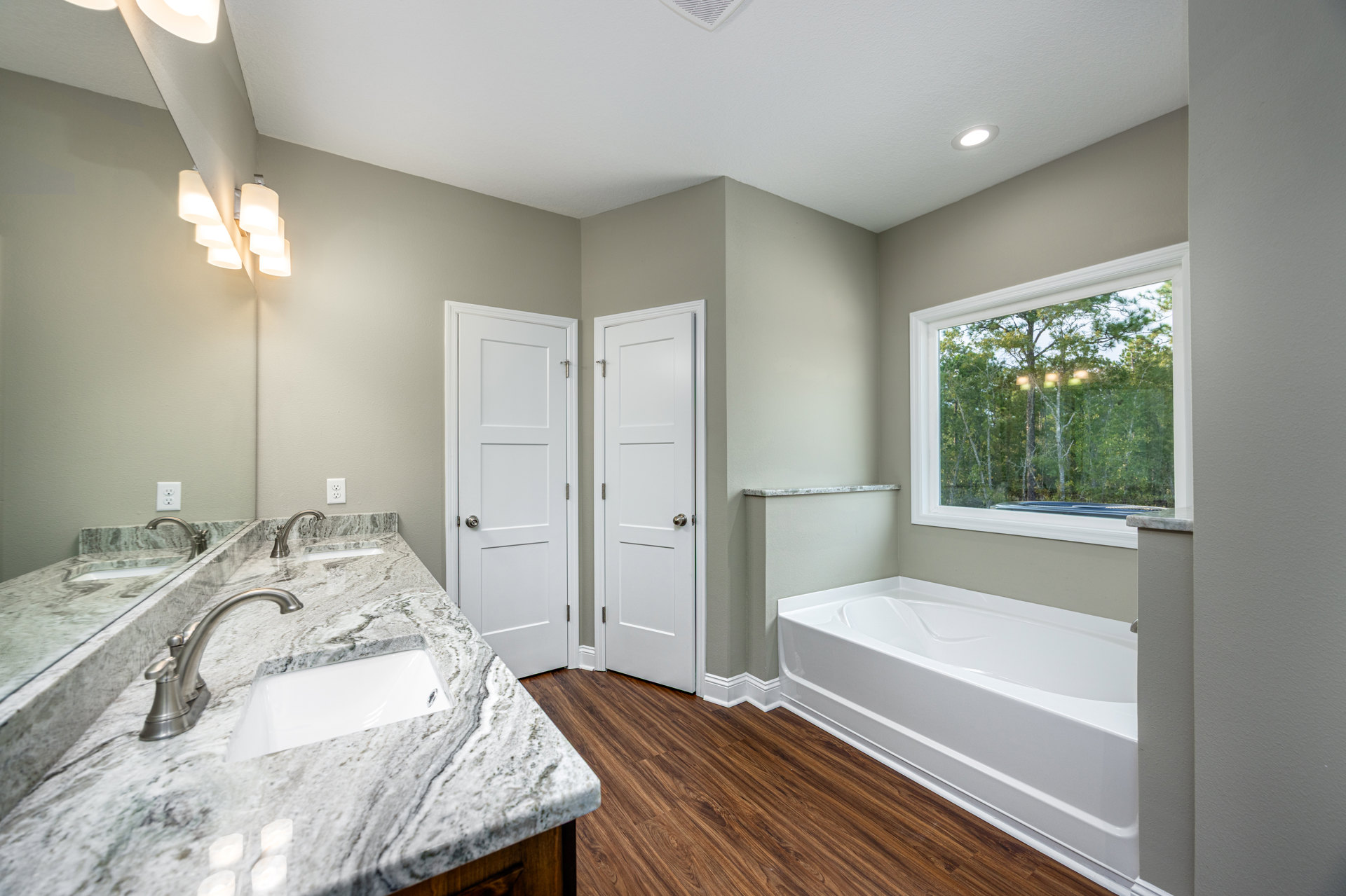 Marble bathroom countertop with undermount sink and chrome faucet, white freestanding bathtub near window overlooking trees, white door with silver knob, large mirror above vanity