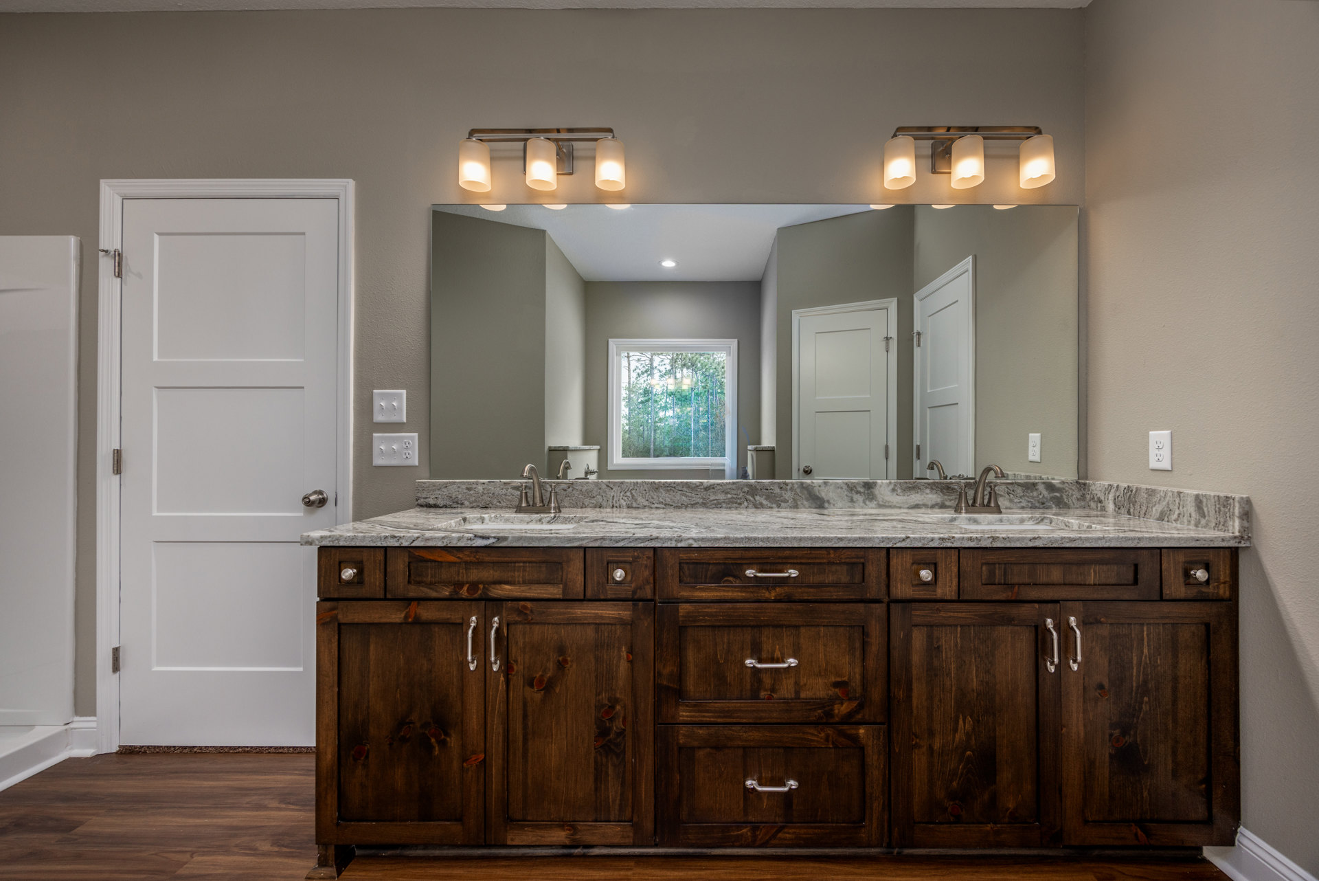 Bathroom with double sink vanity featuring marble countertop and drawers, large mirror above, white door with silver handle, window showing trees outside, light fixture with shade.
