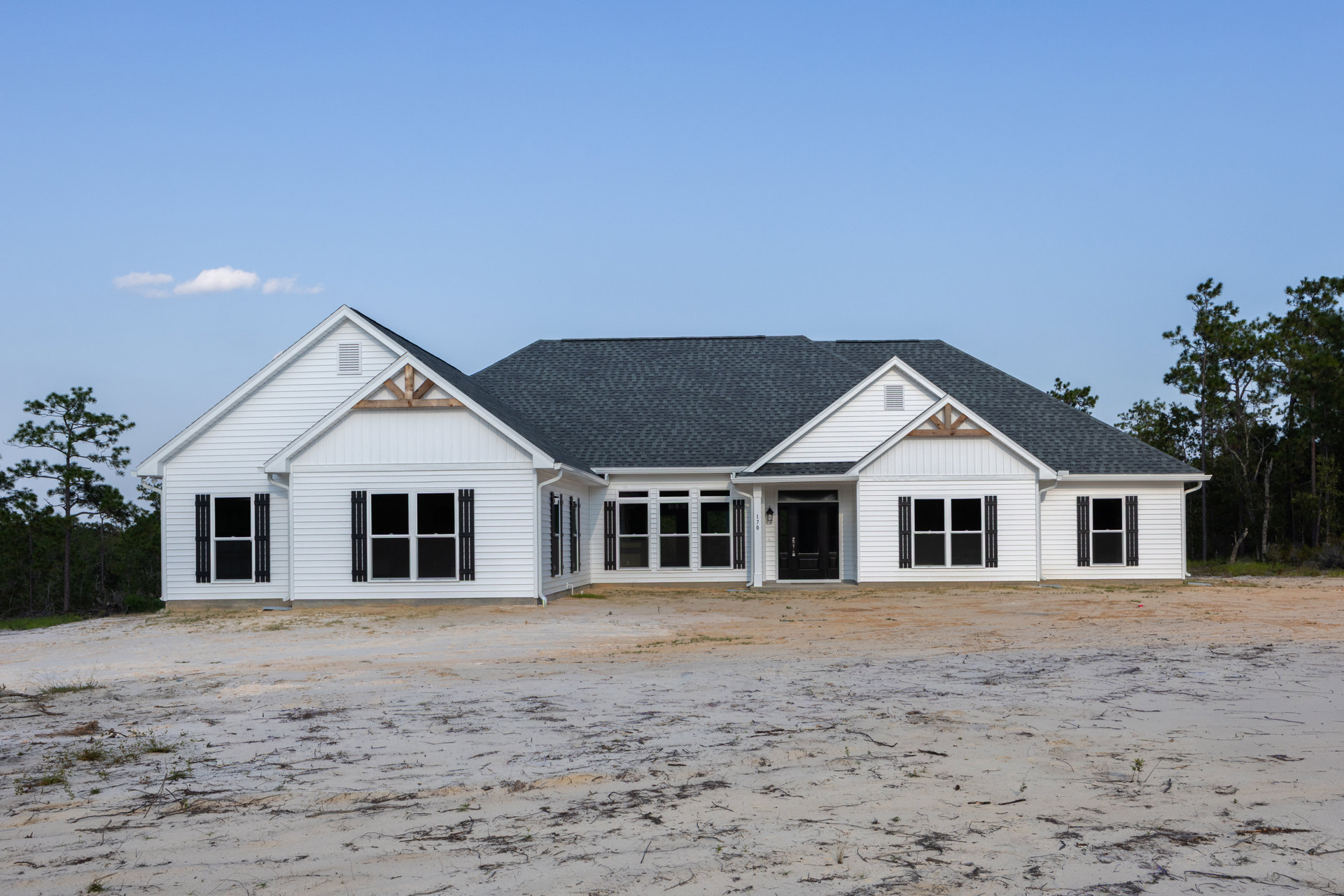 White house with black door and window frames, dirt lot in foreground with scattered sticks, tall tree with bare branches nearby, cloudy sky overhead