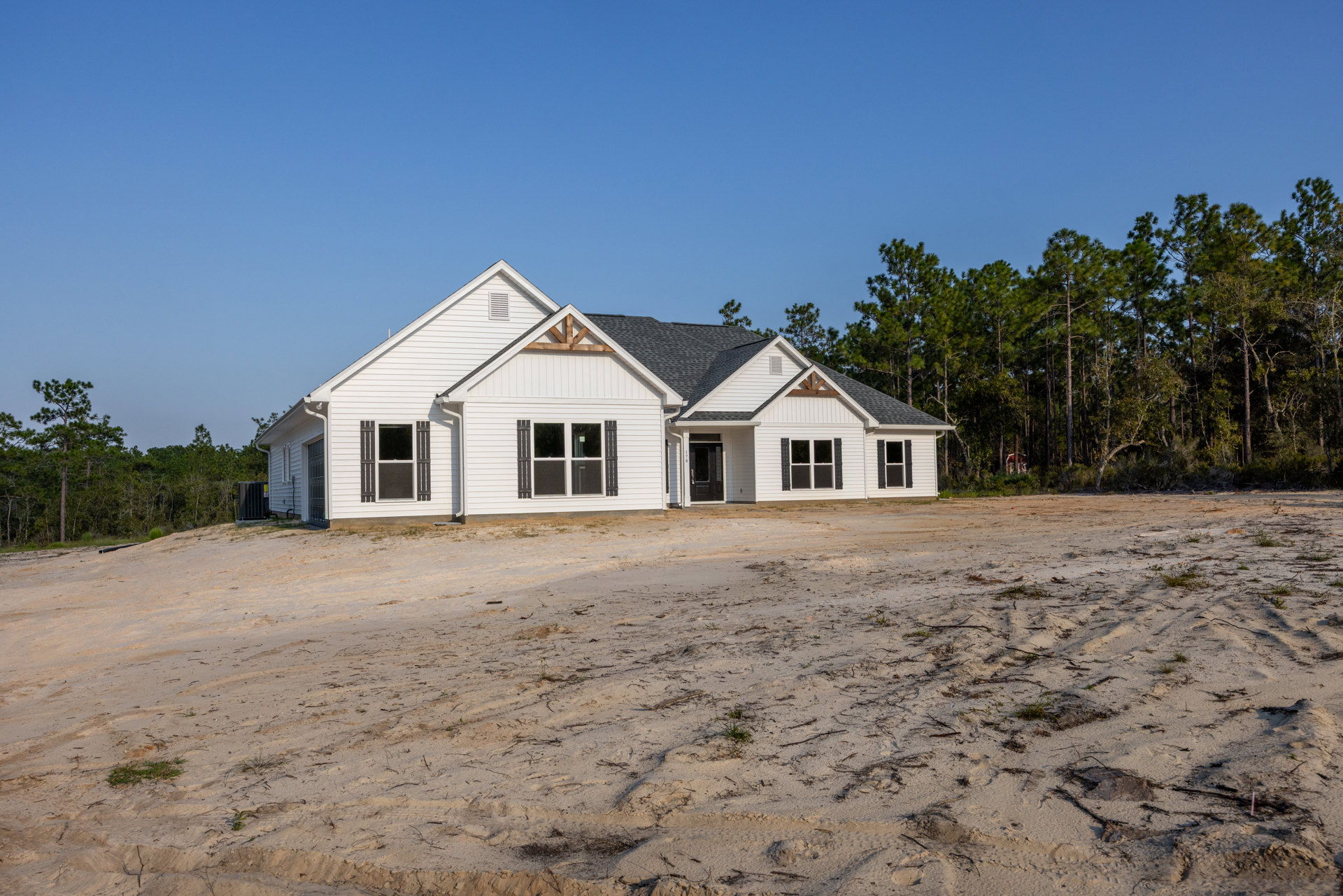 White house with black door and white-framed windows, set against a blue sky, surrounded by trees, with a dirt road and dirt area in the foreground
