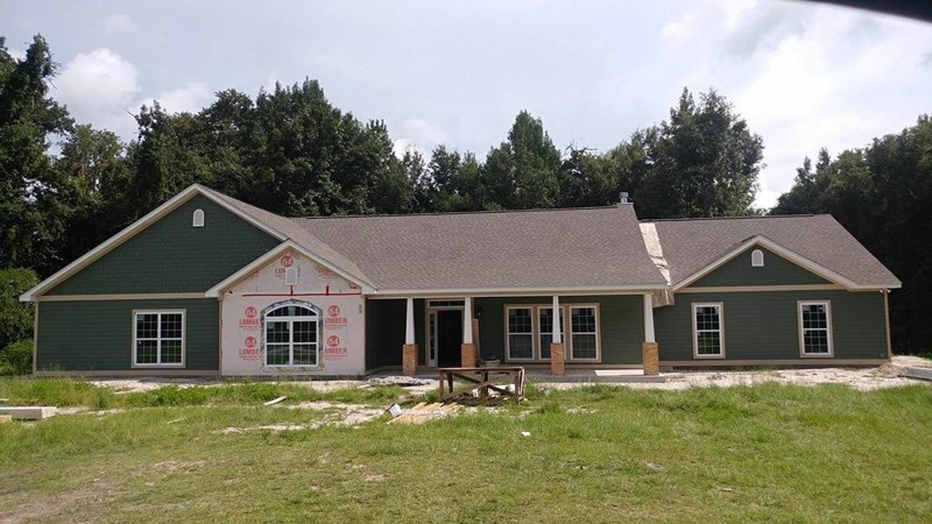 White-walled house with green metal roof, front window, American flag mounted near entrance, wooden fence in foreground, manicured grass lawn, trees and cloudy sky in background