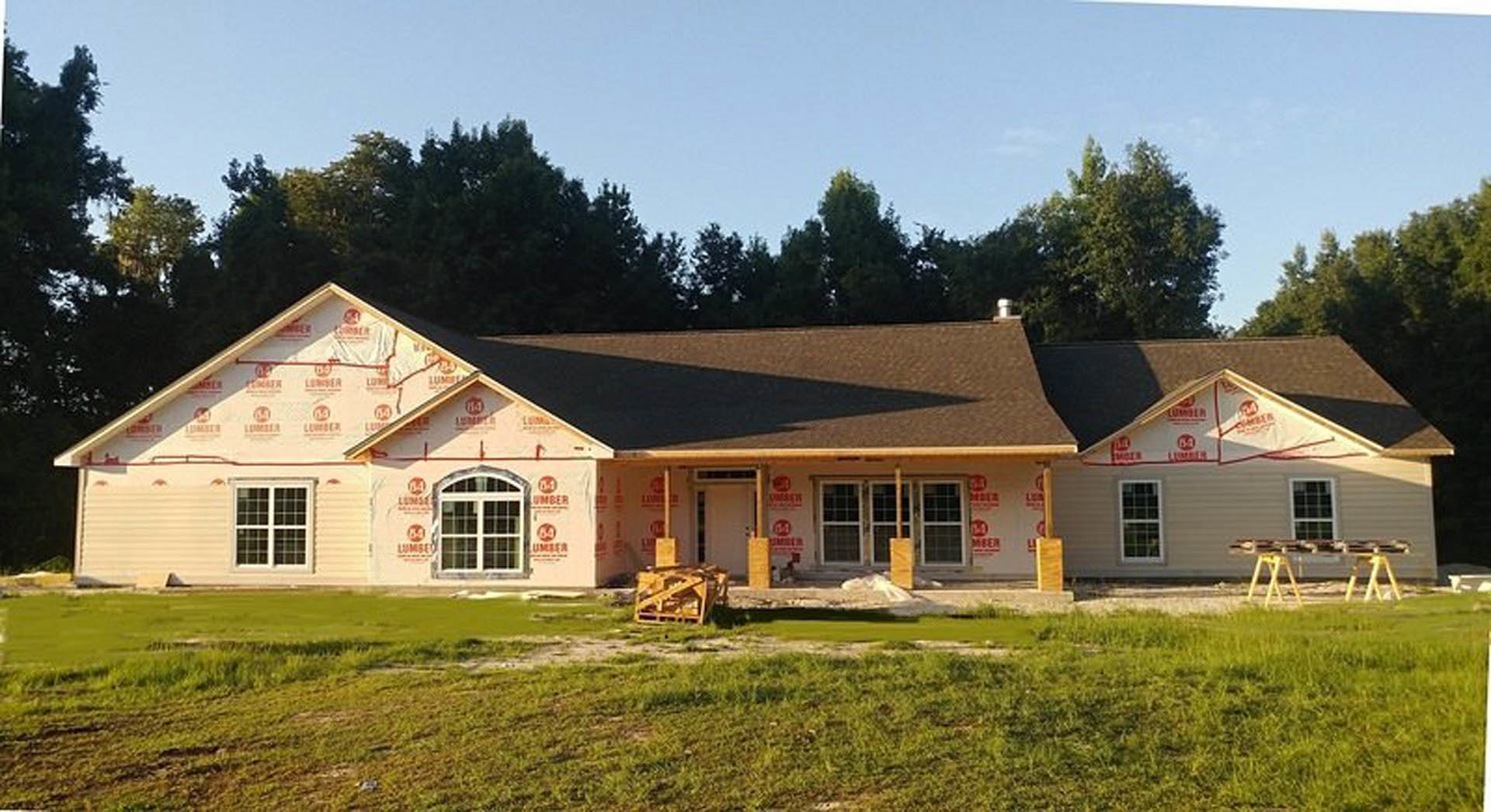 White farmhouse with black-framed windows, green lawn in front, mature trees in background, gabled roof, and covered porch.