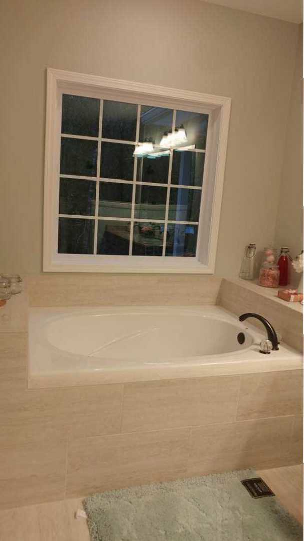 White freestanding bathtub with chrome faucet beside a large window, light reflecting on tiled walls, partial carpet visible in foreground