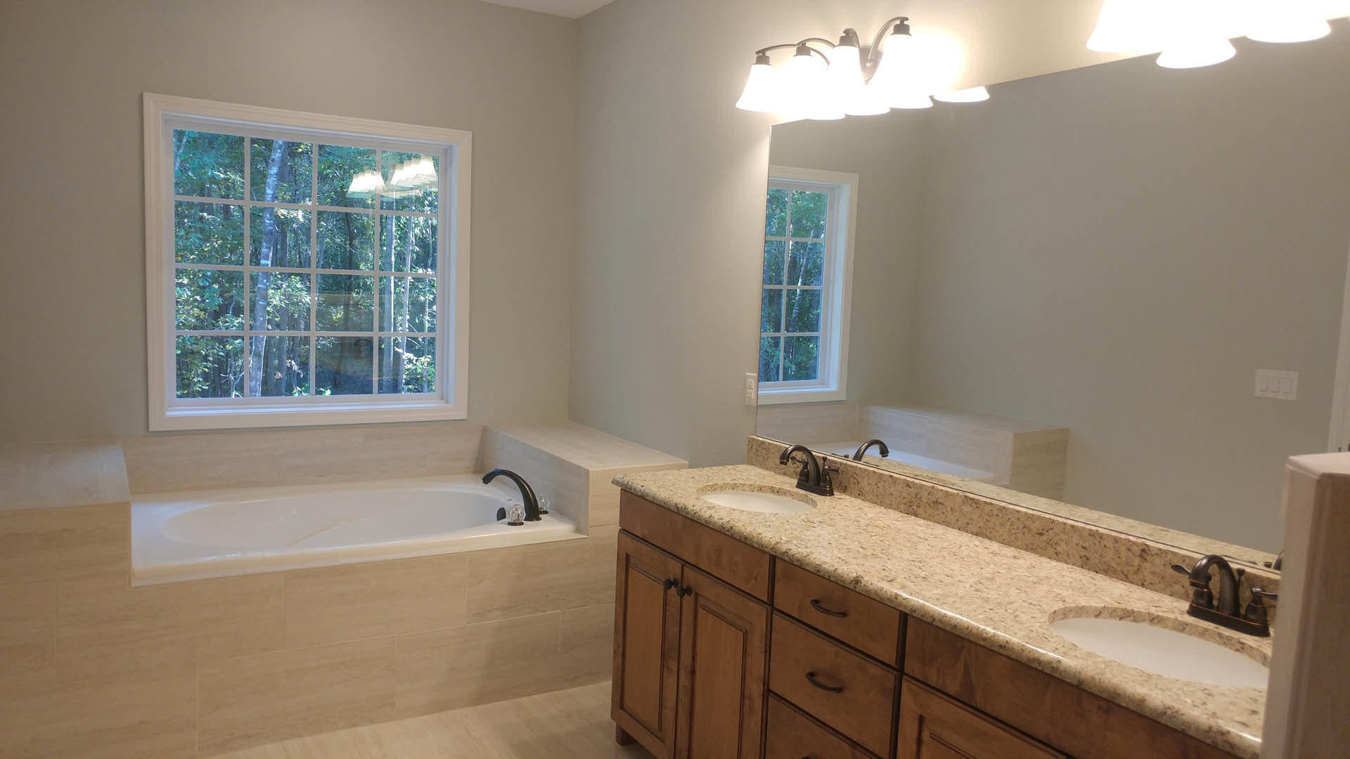 Bright bathroom featuring a freestanding tub, dual sinks set in a white countertop, tile flooring, cabinetry with drawers, and large windows overlooking a leafy tree.